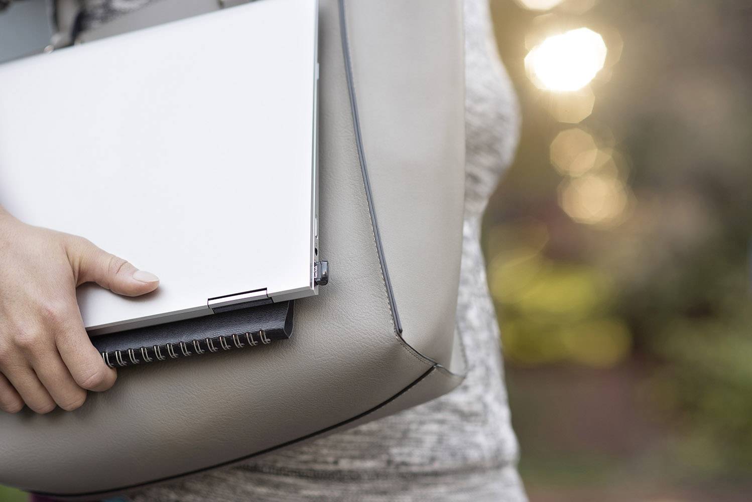 Een persoon draagt een lichtgrijze tas met een laptop en een notitieboek, buiten bij zonsondergang.