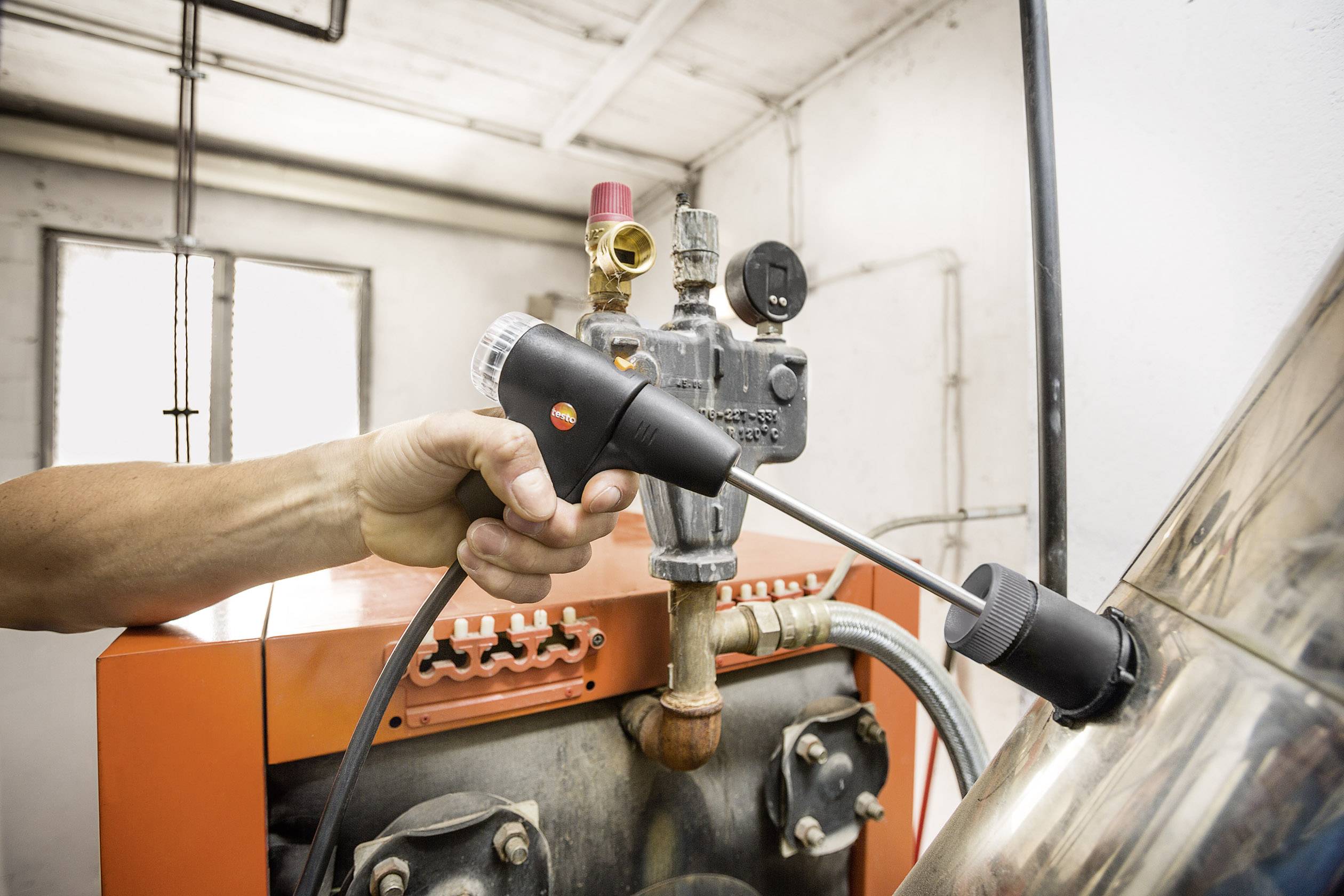 Een hand houdt een meetinstrument tegen een pijp in een industriële ruimte. Het apparaat zou kunnen worden gebruikt voor het controleren van druk of temperatuur.