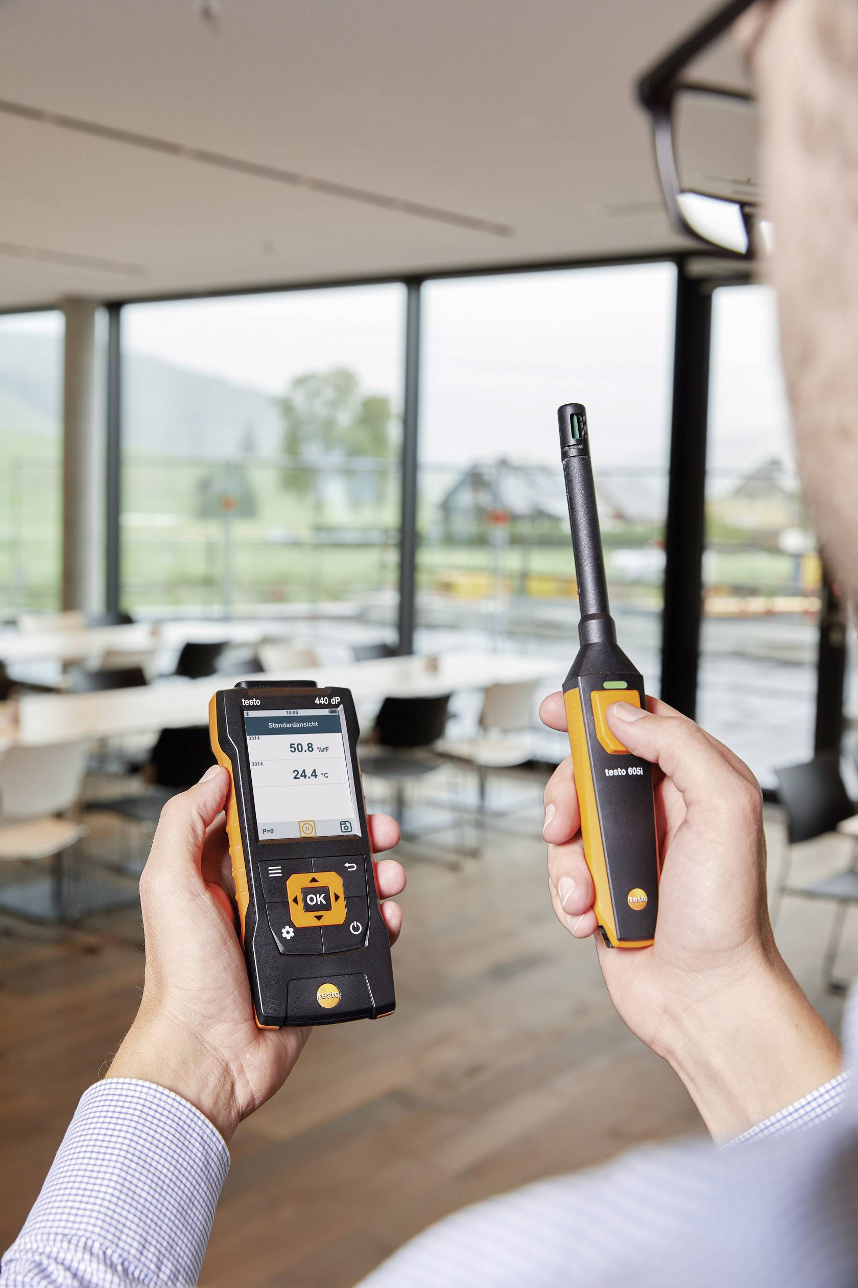 Een man houdt een meetinstrument en een sensor vast in een moderne ruimte met grote ramen en uitzicht op een bergachtig landschap.