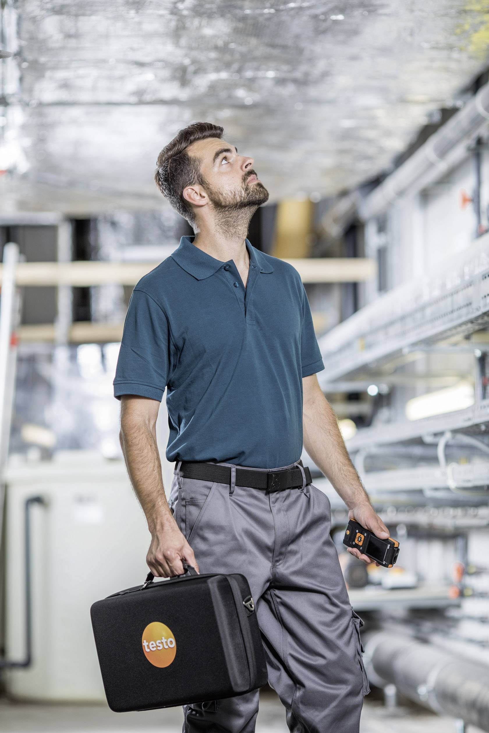 Technicus inspecteert een industriële installatie, draagt een poloshirt en houdt een gereedschapstas en meetapparaat vast.