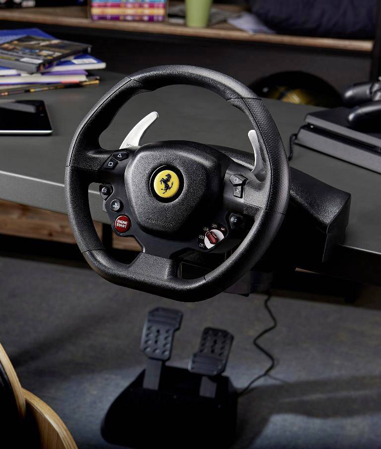 Stuurwiel met Ferrari-logo, gemonteerd op een tafel, met pedaalsysteem eronder. Op de achtergrond zijn boeken en een helm te zien.