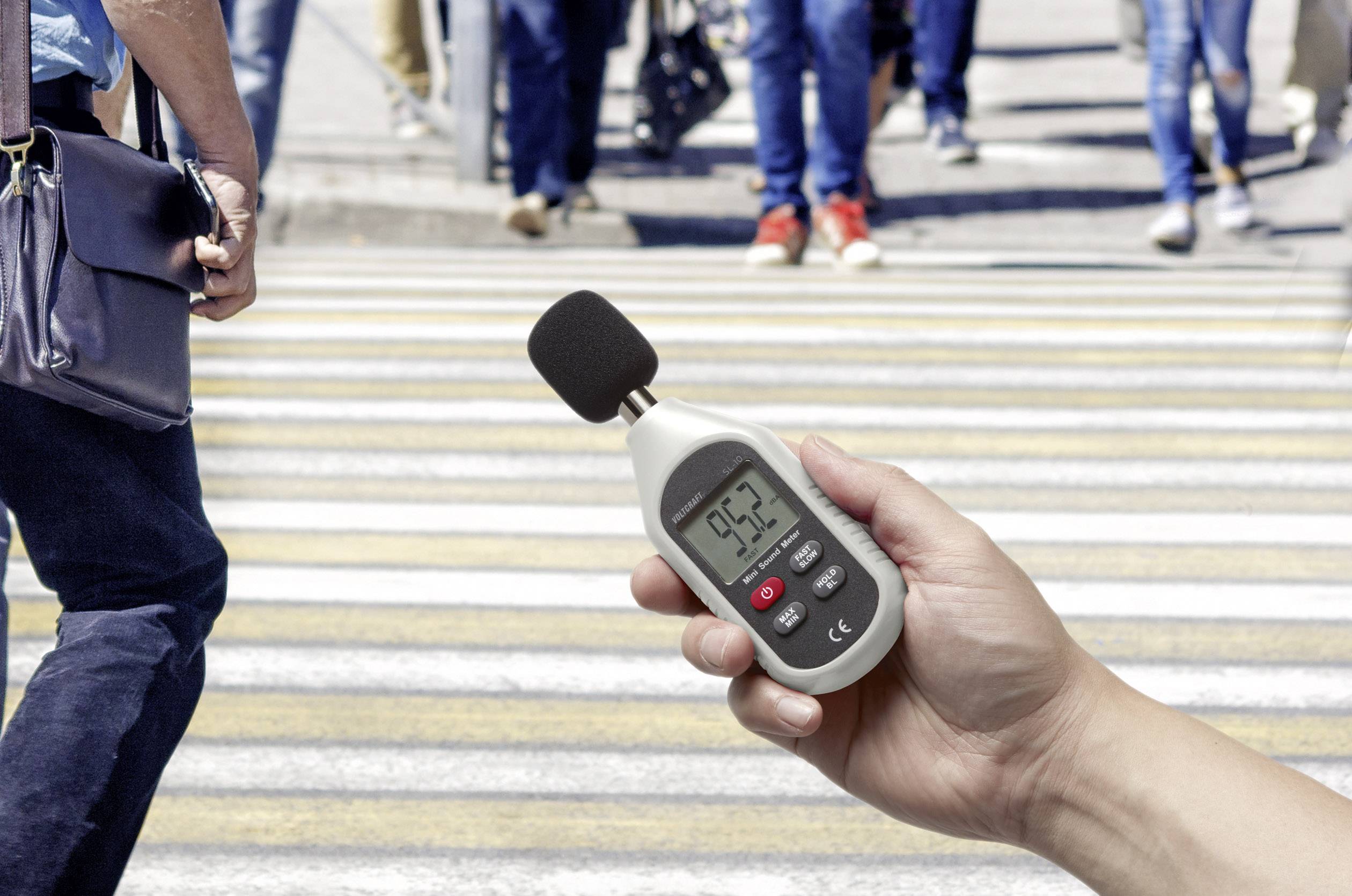 Een hand houdt een geluidsniveaumeter vast die 95,3 decibel aangeeft. Op de achtergrond steken mensen een zebrapad over.