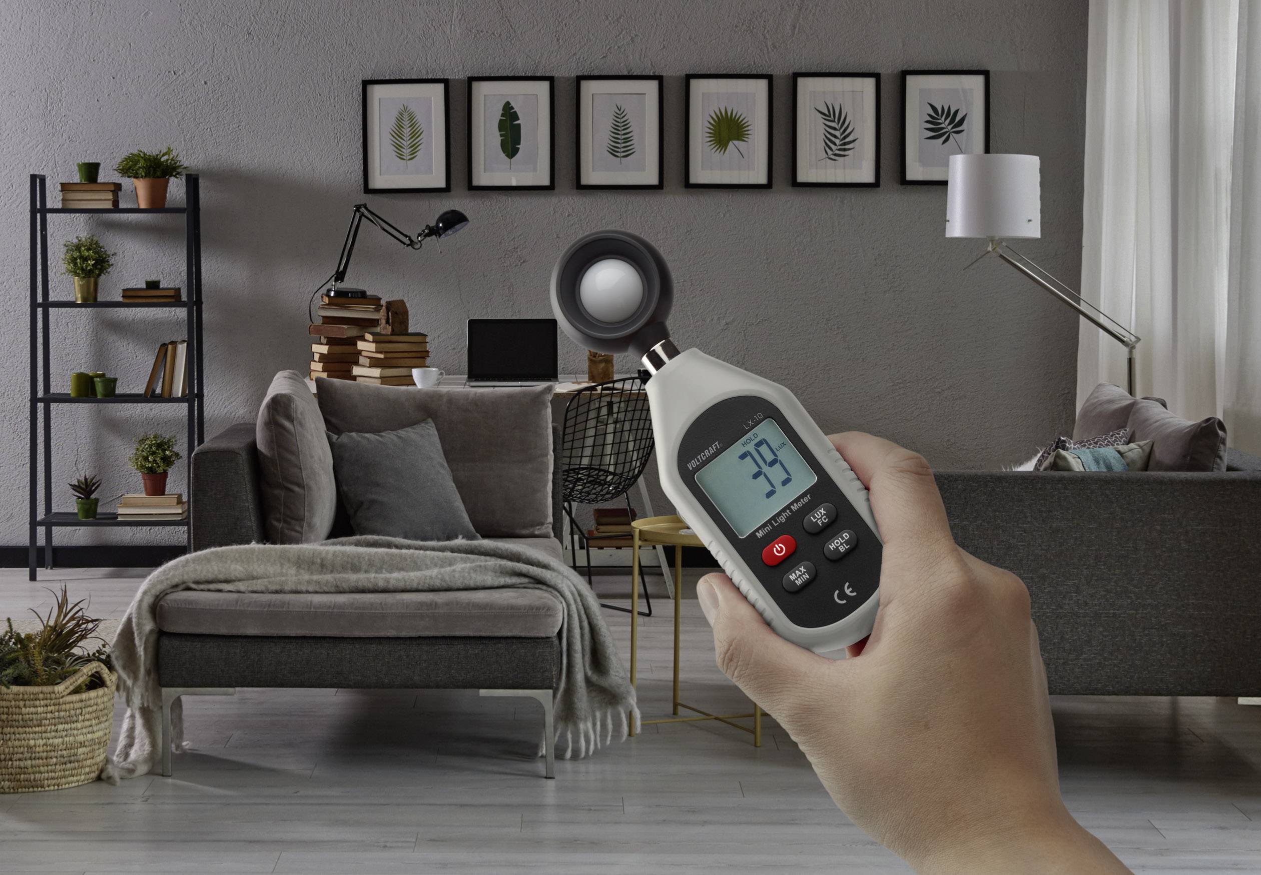 Een handapparaat toont '39.8', vastgehouden in een modern woonkamer met een grijs bankstel, planten, planken en een bureau op de achtergrond.