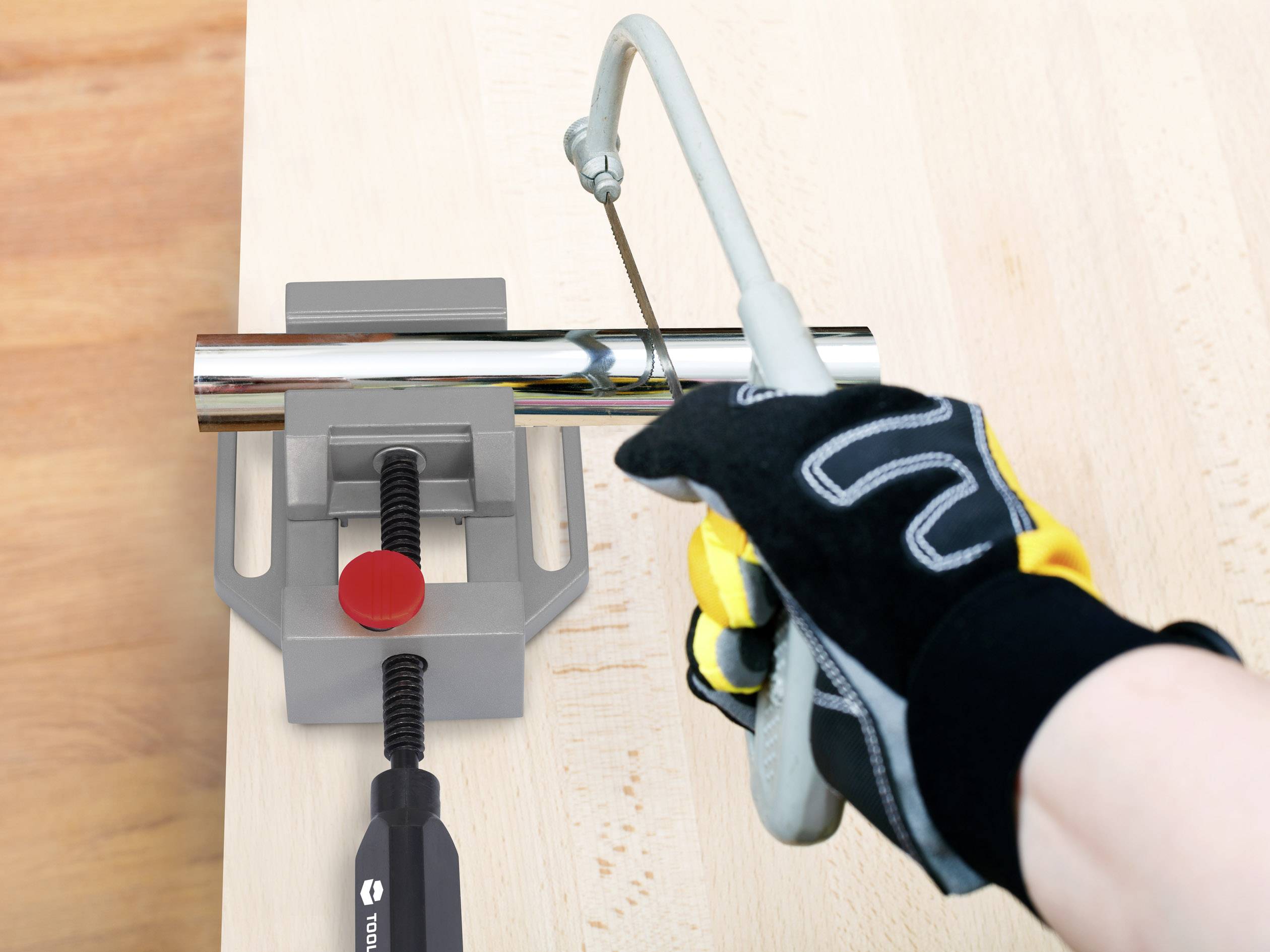 Een hand die metalen gereedschapshandschoenen draagt en een metaalzaag vasthoudt. Een metalen buis wordt in een bankschroef geklemd en doorgezaagd.
