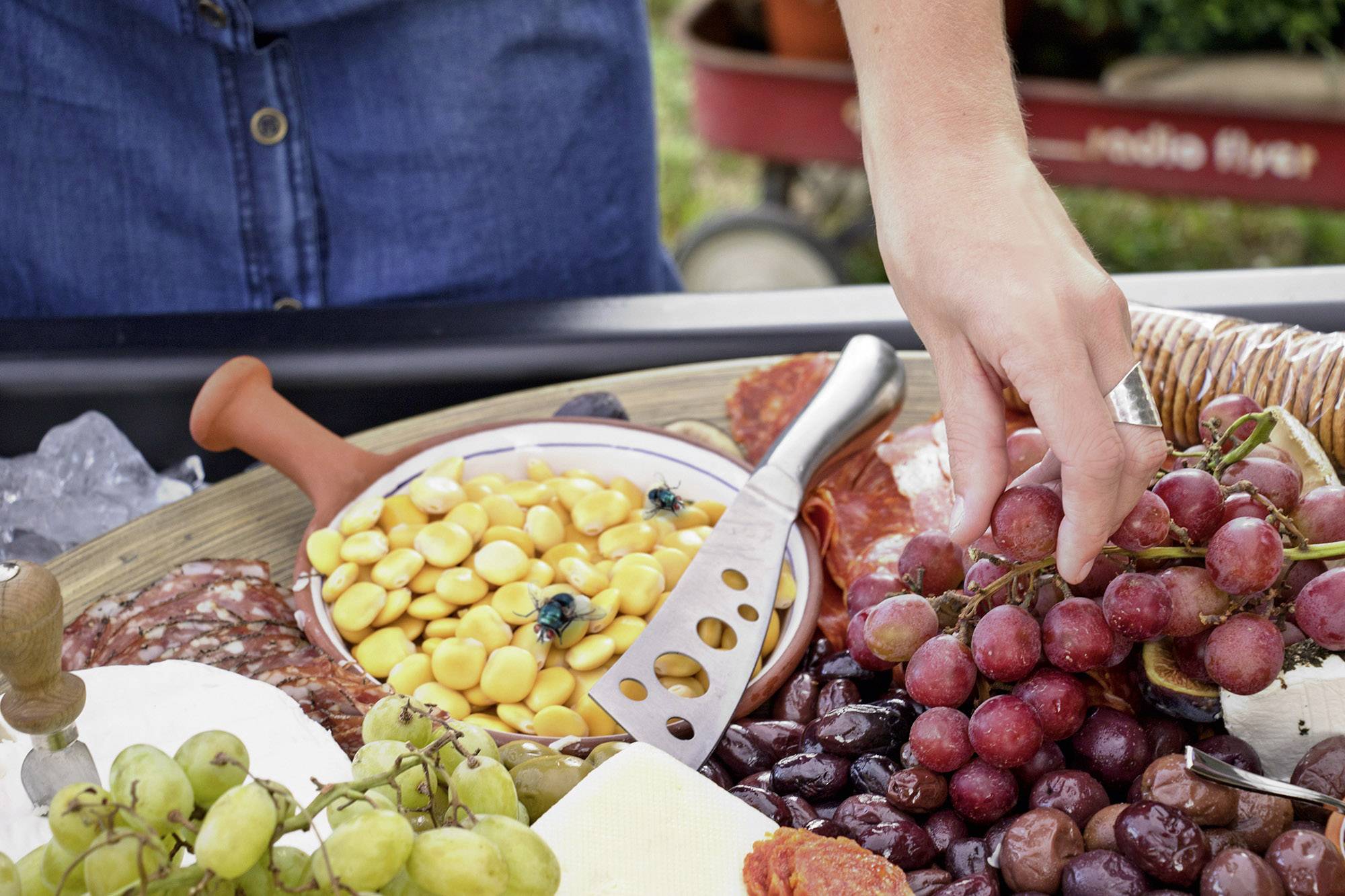 Een hand grijpt naar een bundel rode druiven naast een plateau met verschillende kaassoorten en koude vleeswaren.