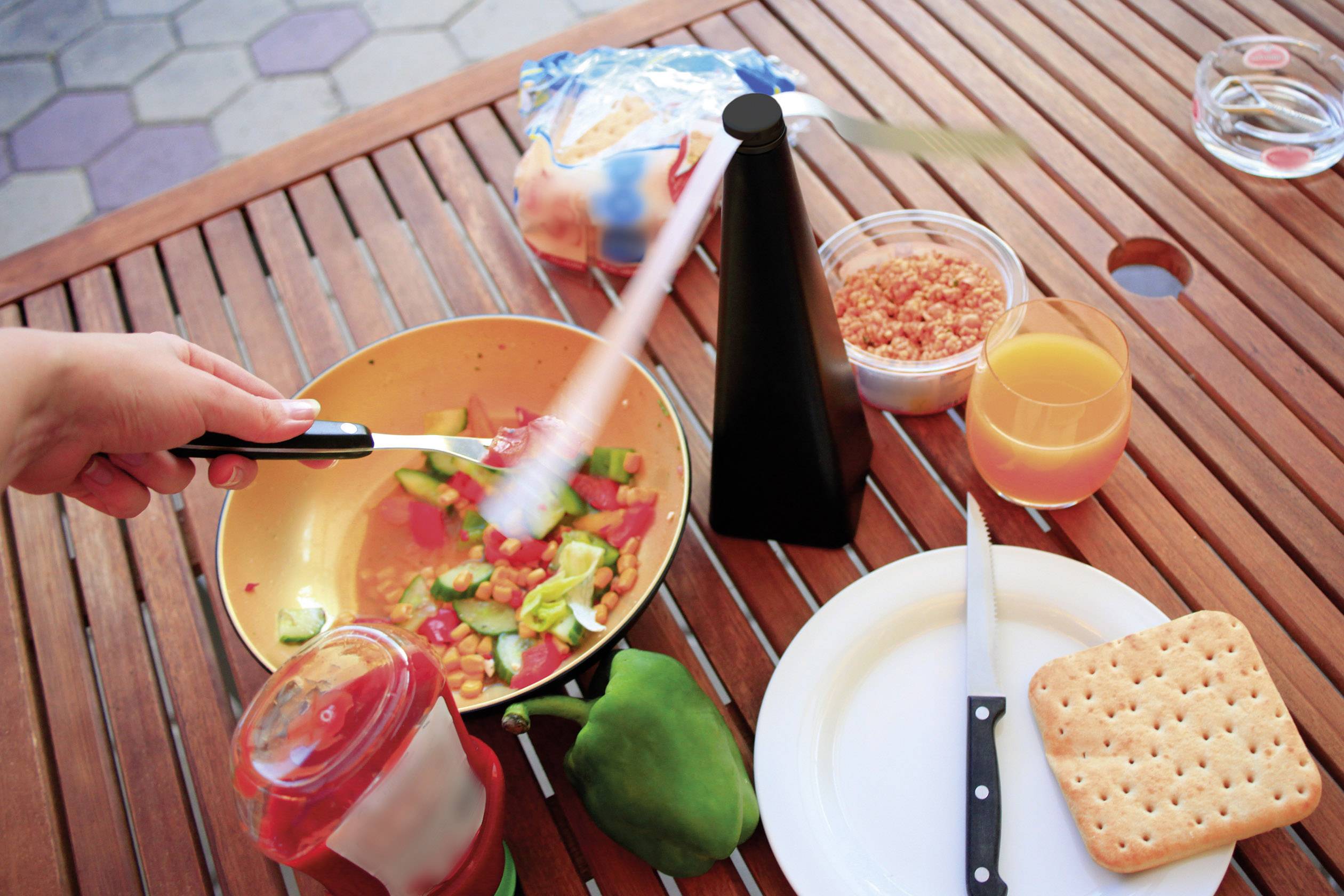 Een tafel met een houten plank, daarop een pan met groenten, een broodtas, sinaasappelsap, een koekjesdoos, een groene paprika en een mes.