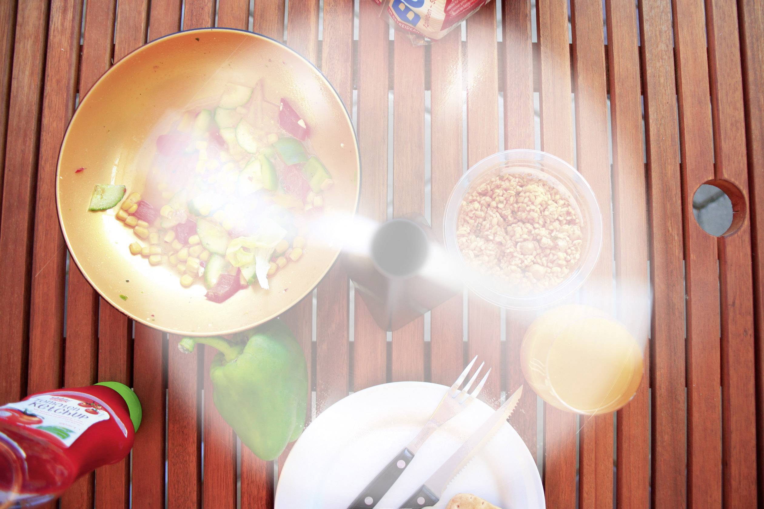 Slaschaal met groenten en ketchupfles op houten tafel, gedeeltelijk verborgen door rotorbladen van een plafondventilator.