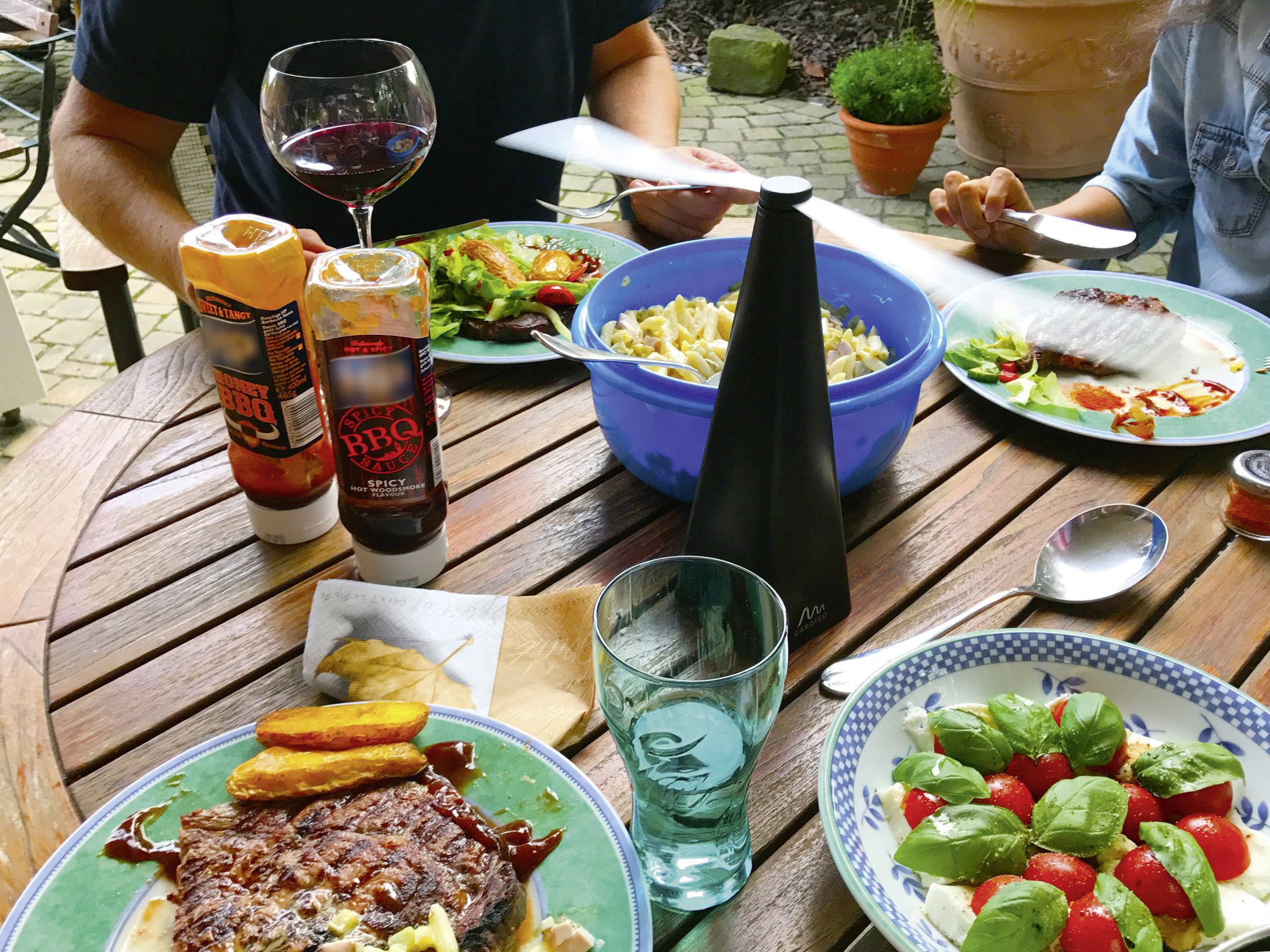 Op een houten tafel buiten staan borden met steak, salade en pasta. Twee personen zitten en eten. Sausflesjes en een wijnglas zijn zichtbaar.