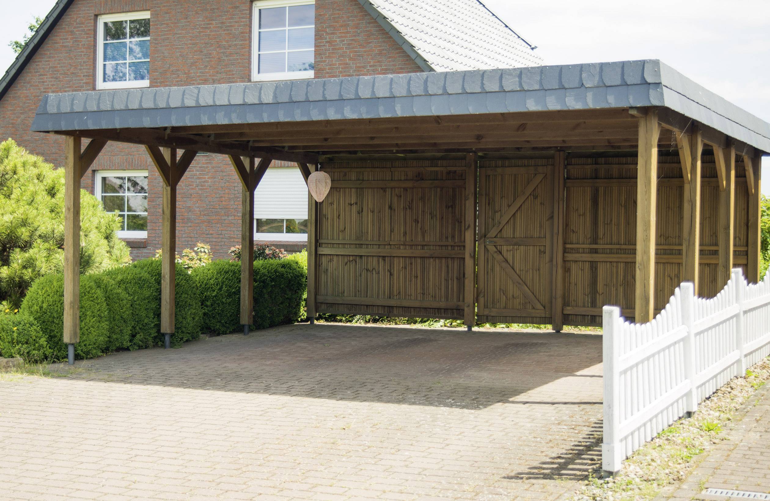 Een houten carport staat voor een bakstenen huis. De grond is bestraat en ernaast staat een witte tuinomheining.