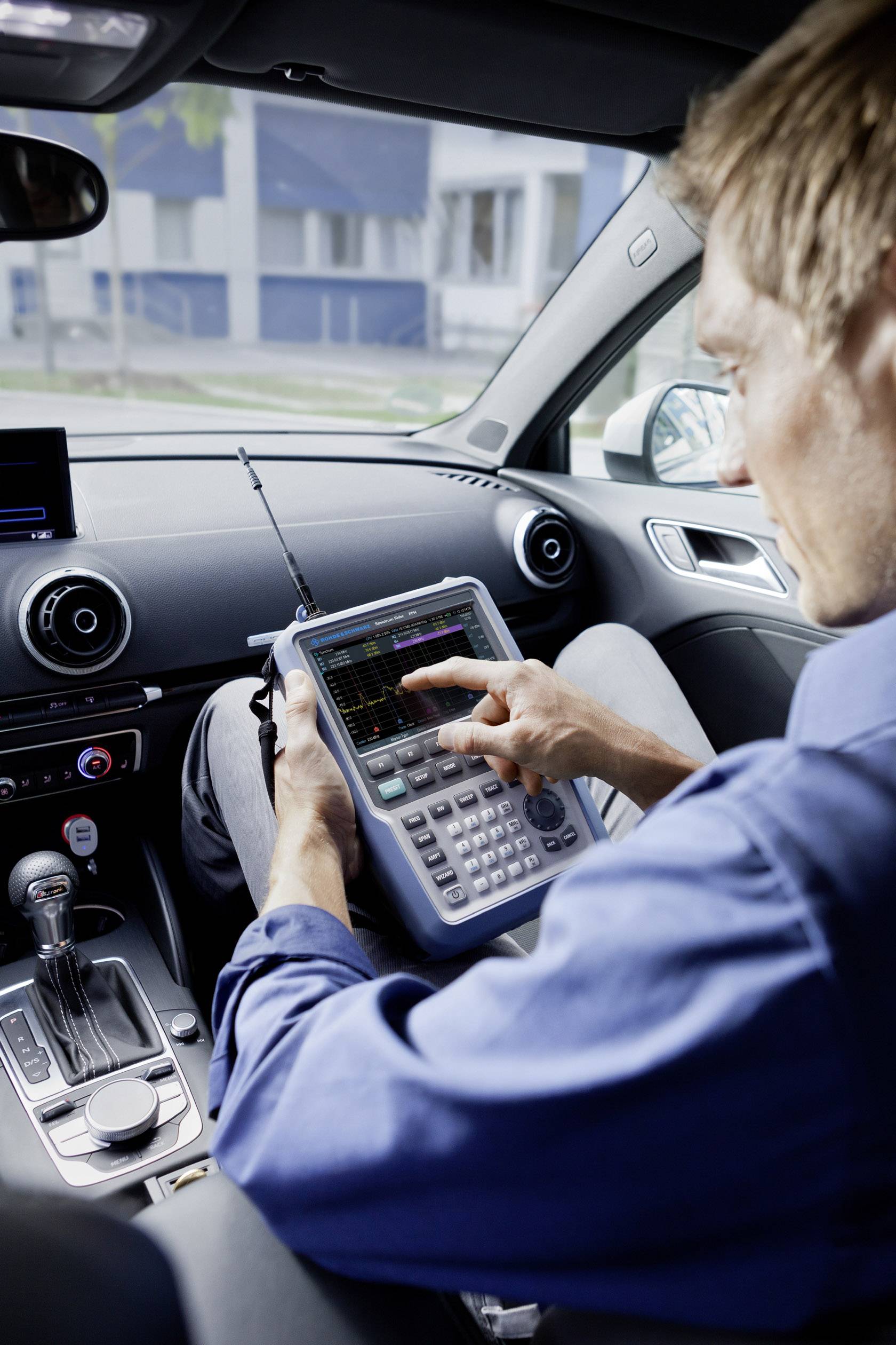 Een man zit in een auto en bedient een draagbaar analyseapparaat. Hij concentreert zich op het display, dat technische gegevens toont.
