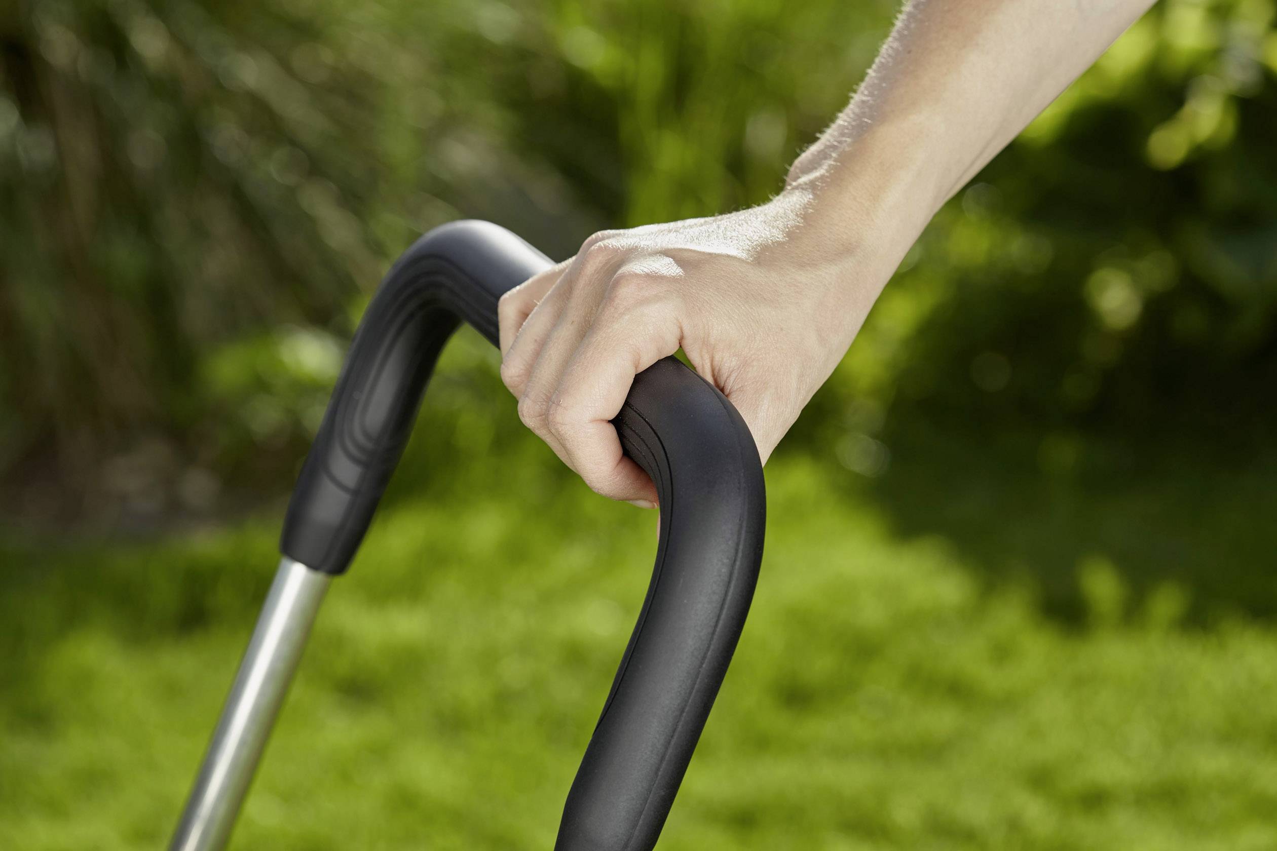 Een hand houdt het handvat van een grasmaaier vast, terwijl groen gazon en planten op de achtergrond te zien zijn.