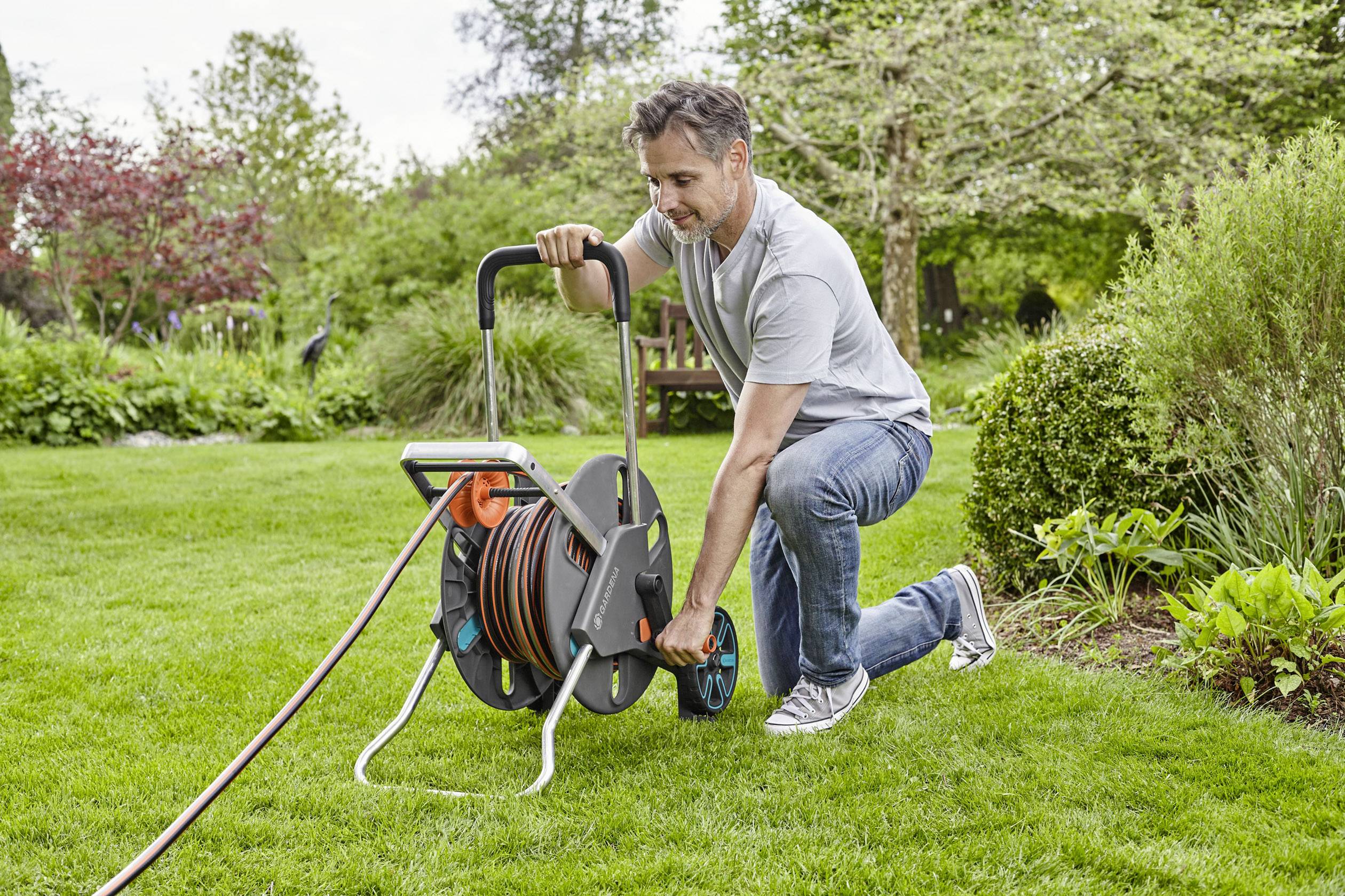 Een man knielt op een grasveld en rolt een tuinslang op een slanghaspel. Op de achtergrond zijn bomen en struiken.