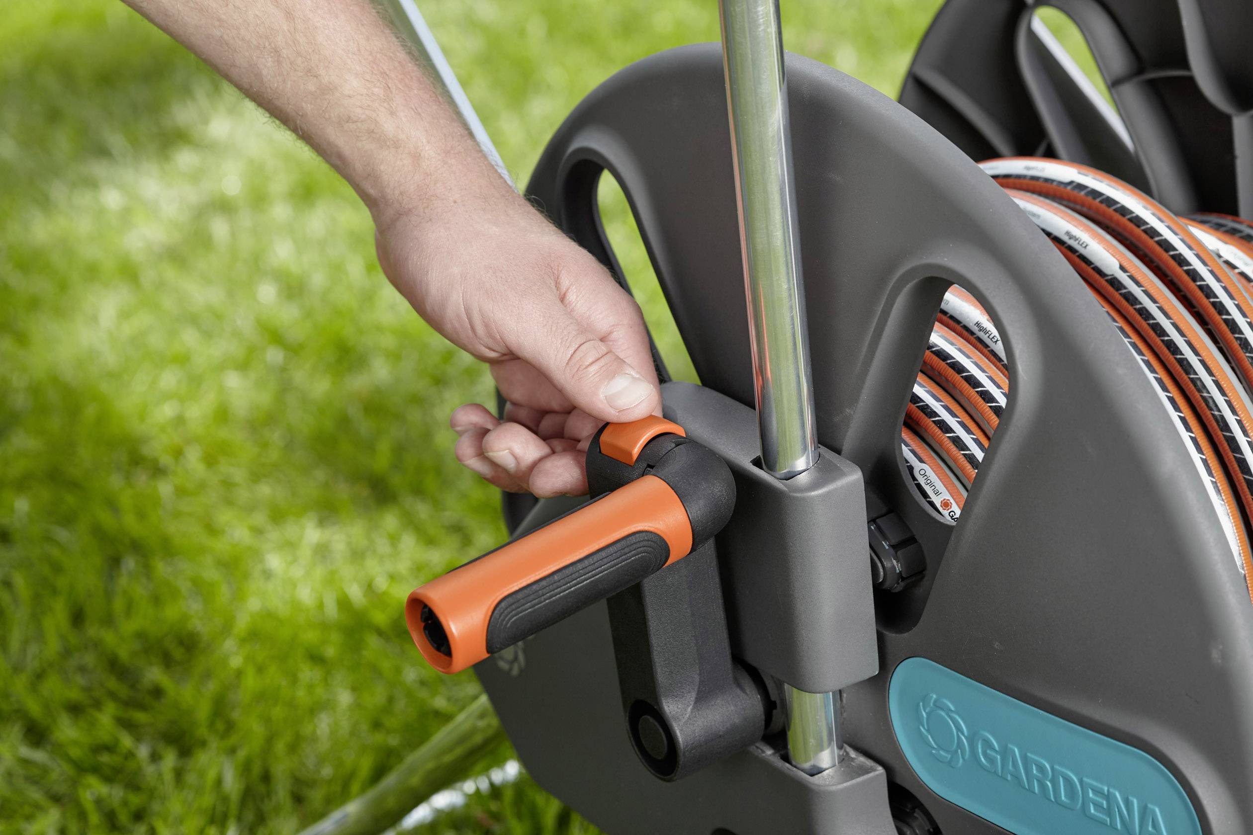 Een hand draait aan een tuinslangoproller op een grasveld. De slangoproller is zwart met oranje accenten.