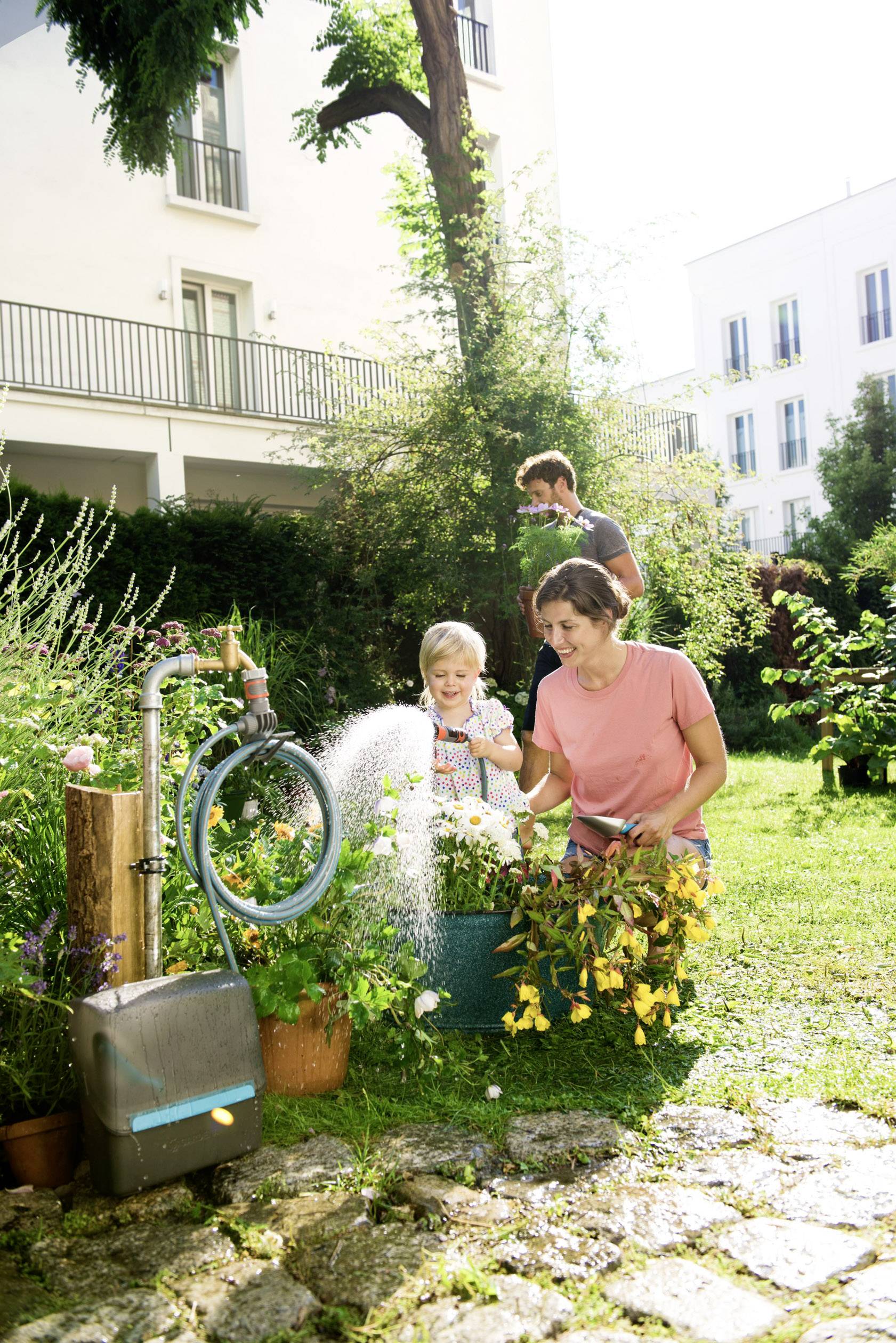 Een familie geeft water aan de tuin: Een vrouw begiet bloemen, een kind houdt de tuinslang vast, terwijl een man op de achtergrond staat.