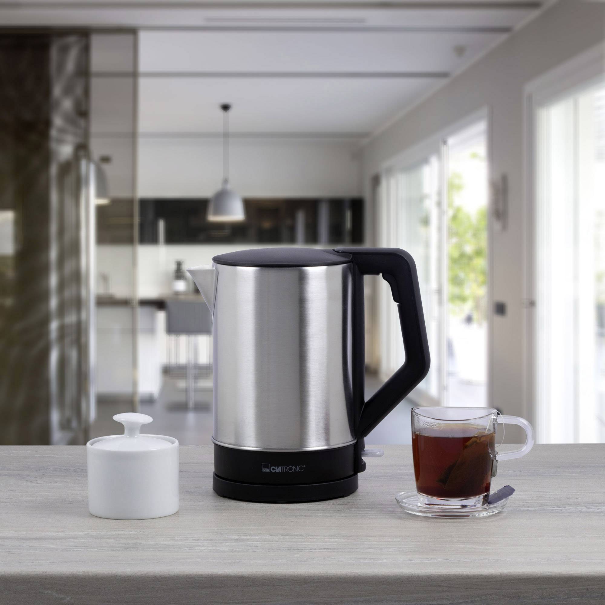 Een roestvrijstalen waterkoker en een kopje thee staan op een tafel in een moderne keuken. Naast de waterkoker staat een suikerdispenser.