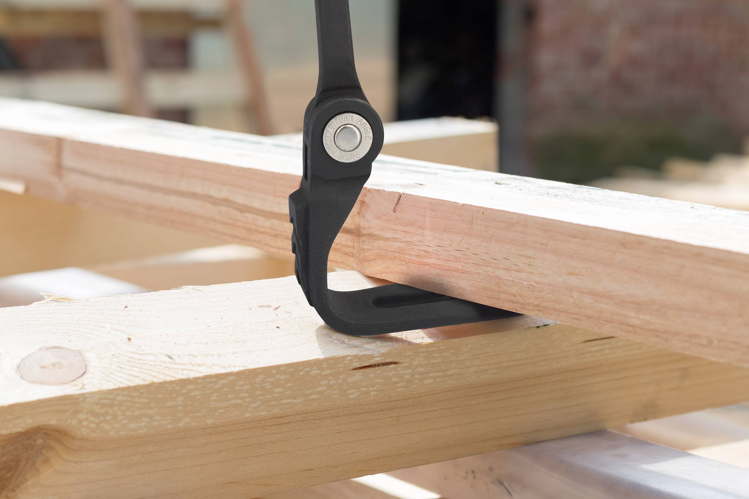 Een zwarte metalen klem houdt twee houten planken bij elkaar die worden voorbereid voor een bouwproject. Op de achtergrond is een wazig werkmilieu zichtbaar.