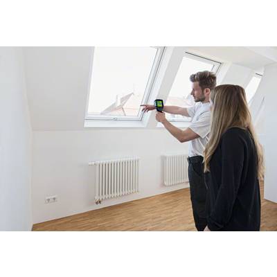 Een persoon toont een warmtebeeldcamera in een ruimte met dakramen en radiatoren om mogelijke warmtelekken te identificeren.