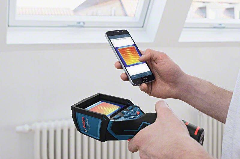 Een persoon houdt een warmtebeeldcamera vast en vergelijkt het beeld op het apparaat met een display op een smartphone. De achtergrond toont een raam.