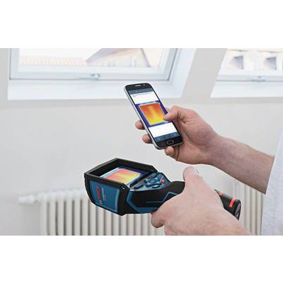 Een persoon houdt een warmtebeeldcamera vast en vergelijkt het beeld op het apparaat met een display op een smartphone. De achtergrond toont een raam.