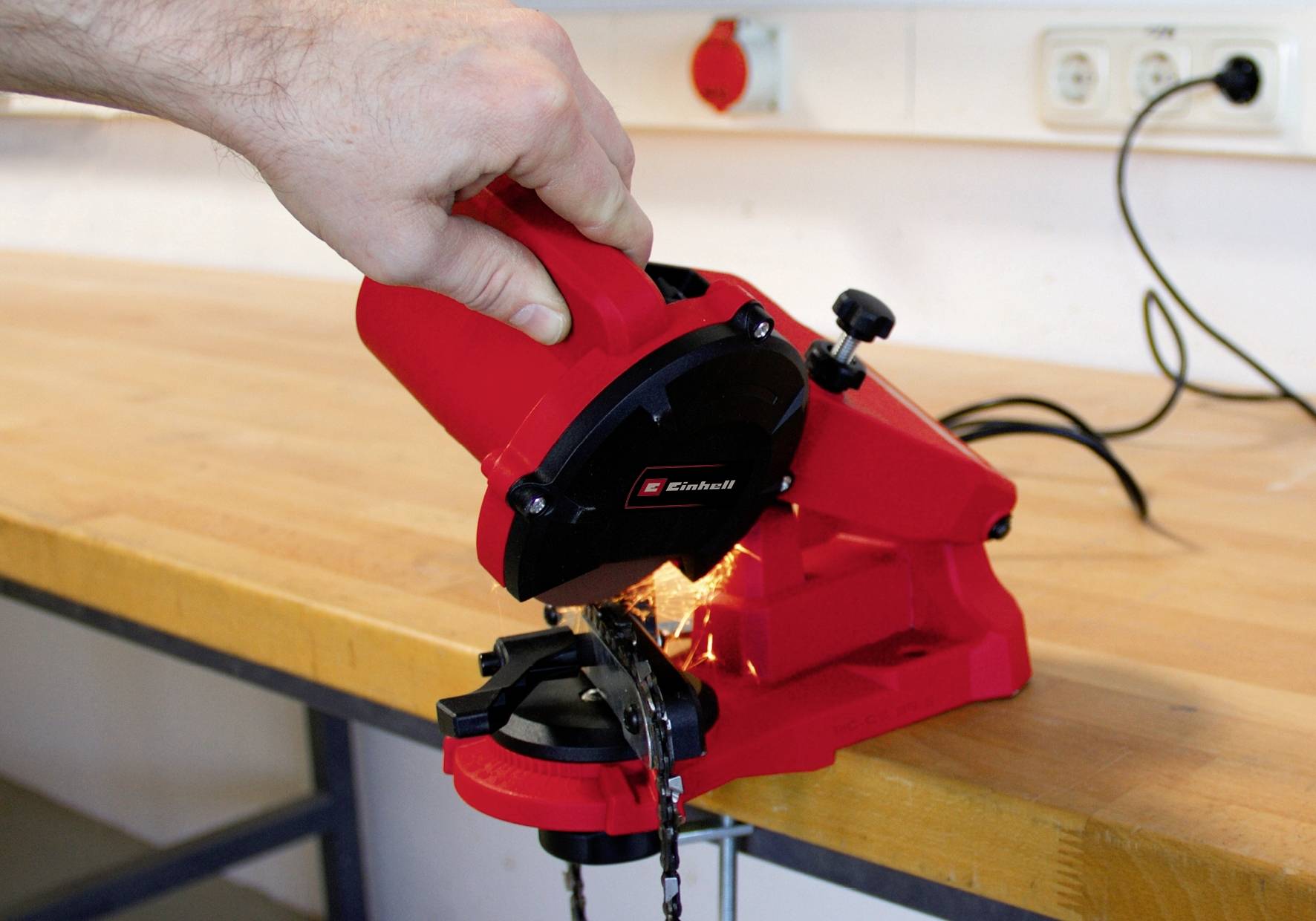 Een hand bedient een rode kettingscherper op een werkbank. Vonken spatten rond terwijl de ketting wordt geslepen.