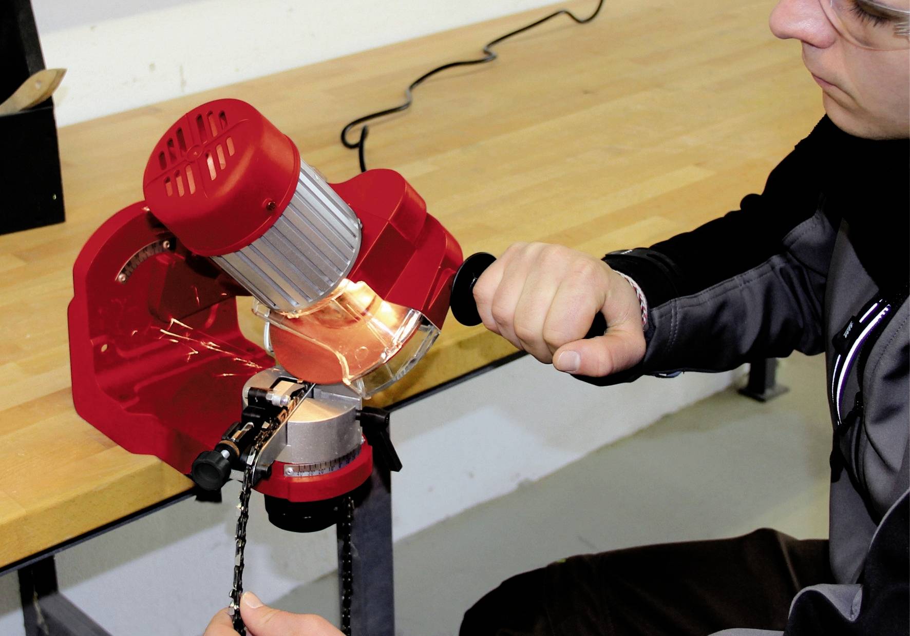 Een persoon gebruikt een rode slijpmachine op een werkbank om een ketting te slijpen. De persoon houdt de ketting met één hand vast.
