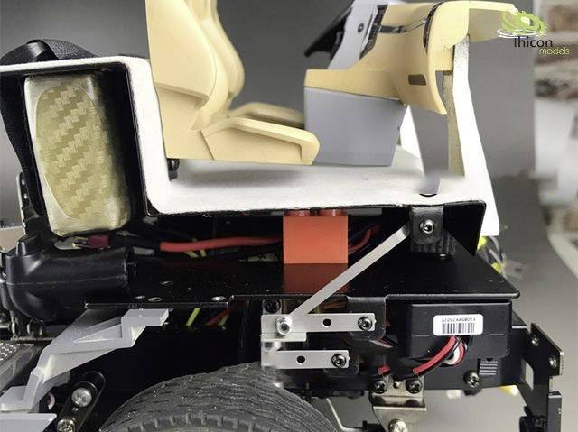 Een modelvoertuigchassis met zichtbare vering en wielophanging. Twee beige stoelen, gedeeltelijk bedekt, op de achtergrond.