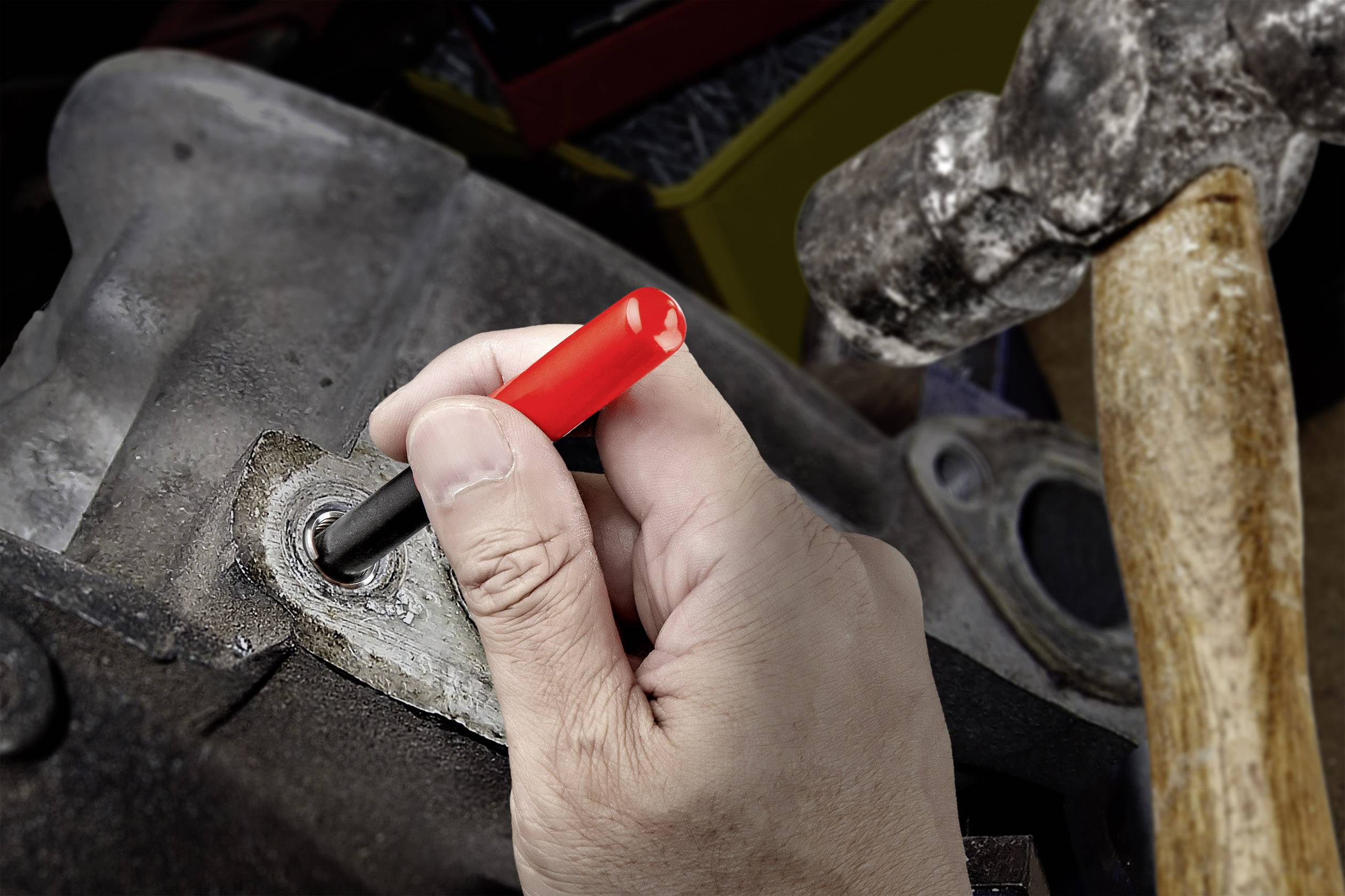 Een hand houdt een rode beitel vast aan een metalen stuk, een hamer is ernaast gepositioneerd, klaar om te slaan.