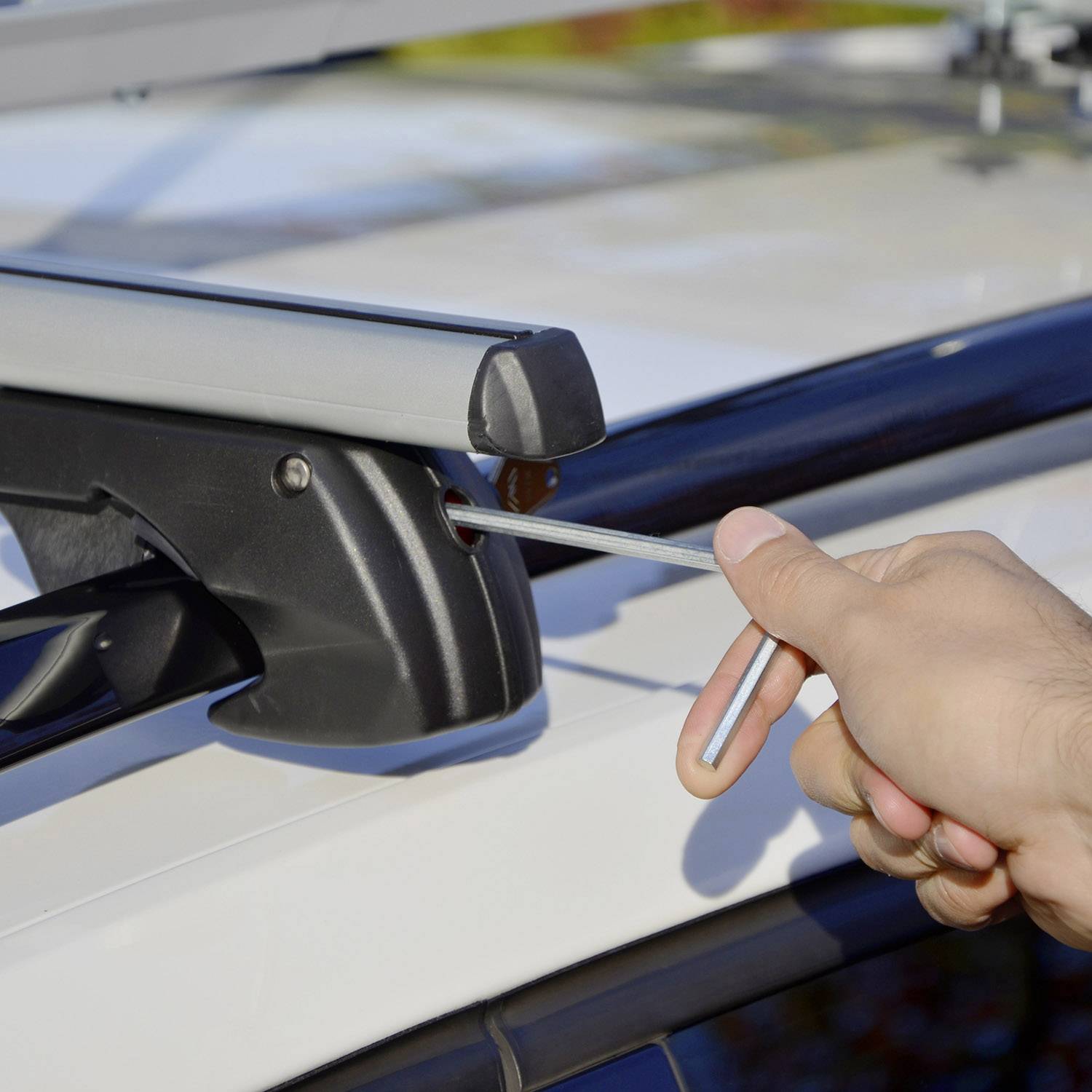 Een hand monteert een dakdrager op het dak van een auto, met een steeksleutel in de hand, op een zonnige dag.
