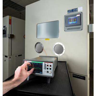 Een hand bedient een elektronisch testapparaat dat is aangesloten op een grote kamer. De kamer heeft een klein display.