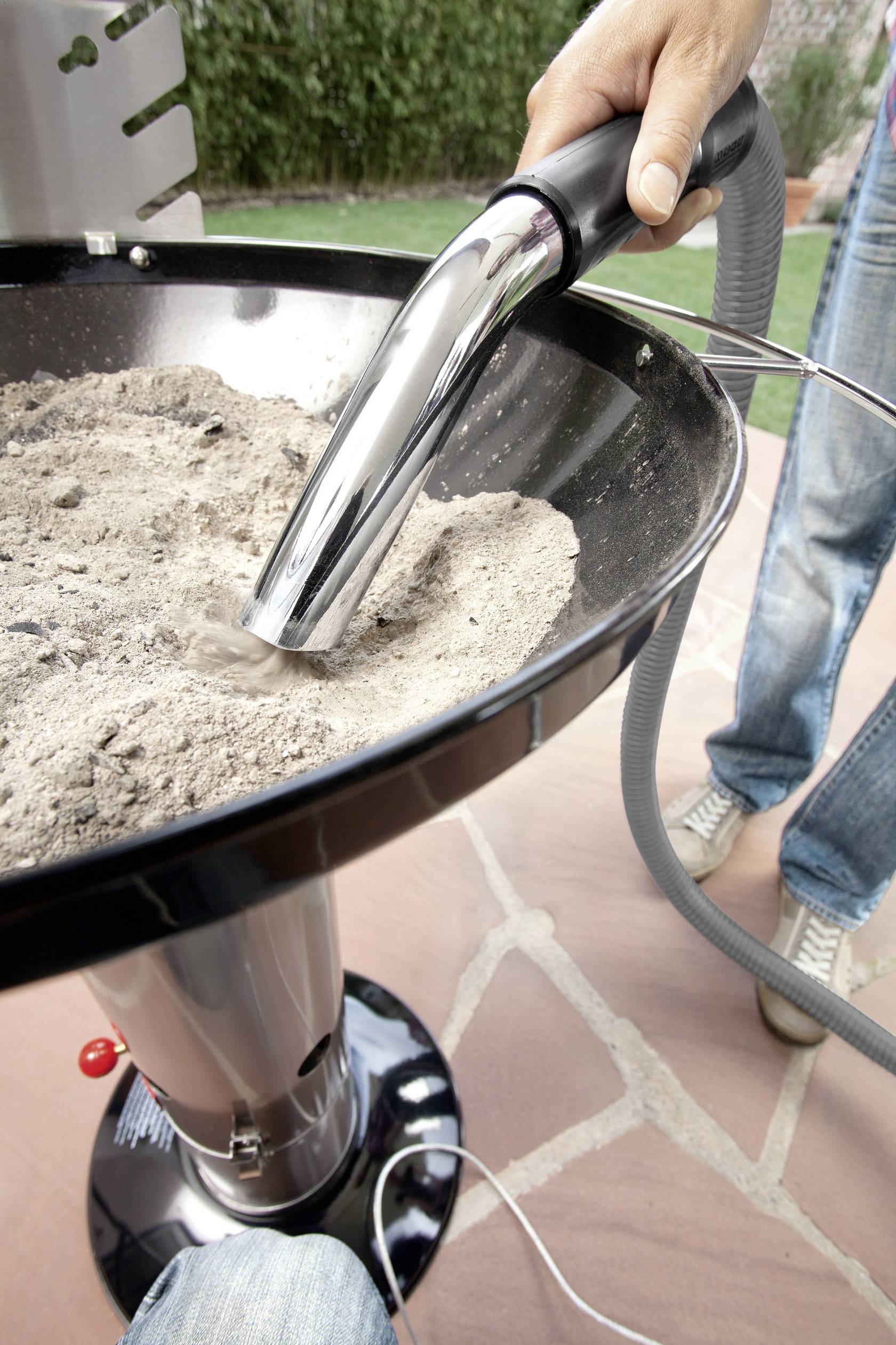 Een persoon zuigt as van een barbecue door een metalen, slankvormige stofzuiger ertegenaan te houden. De locatie is buiten.