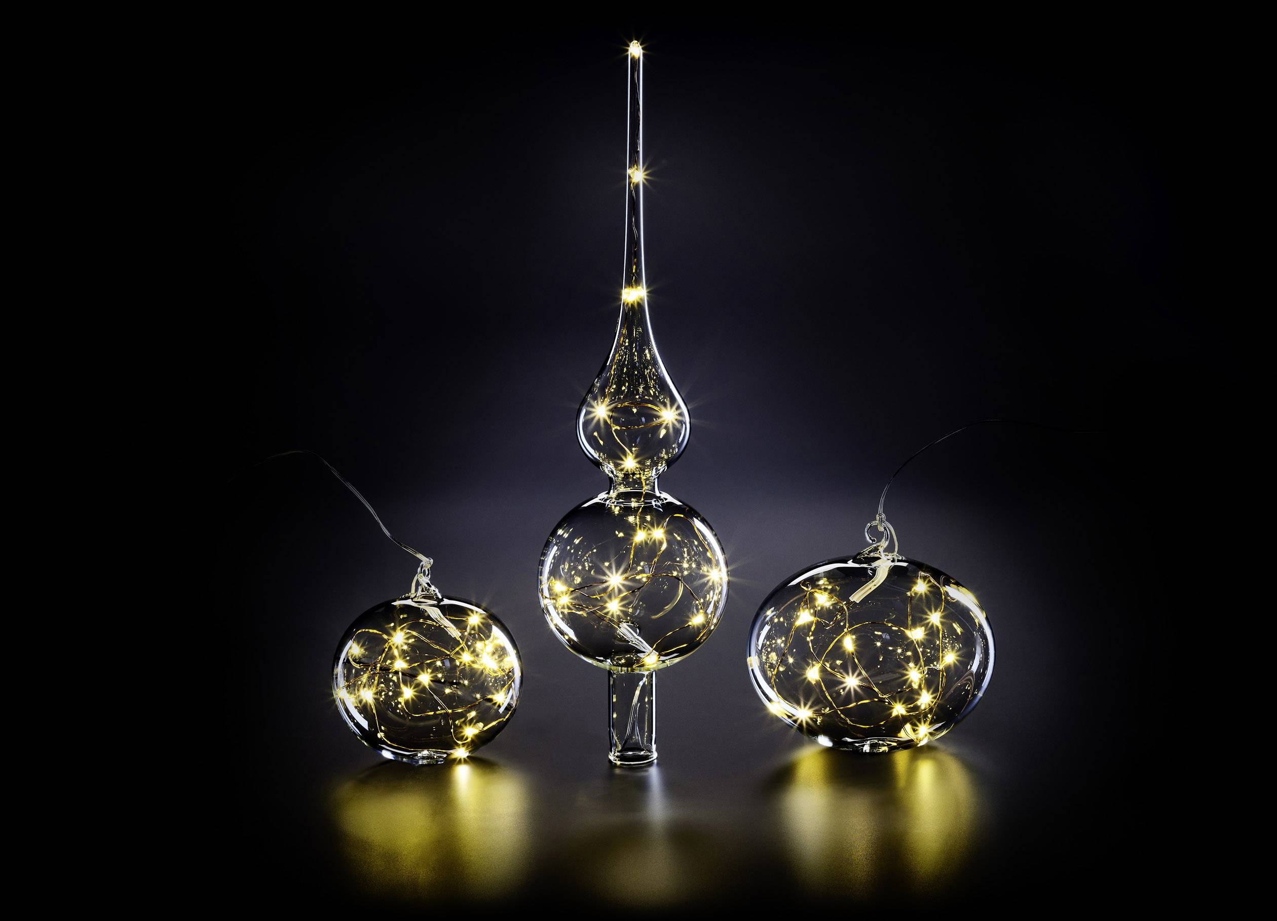 Drie glazen ornamenten op een donkere achtergrond, gevuld met warmwitte snoerverlichting. Het linker en rechter ornament zijn bolvormig; het middelste is hoog en smal.