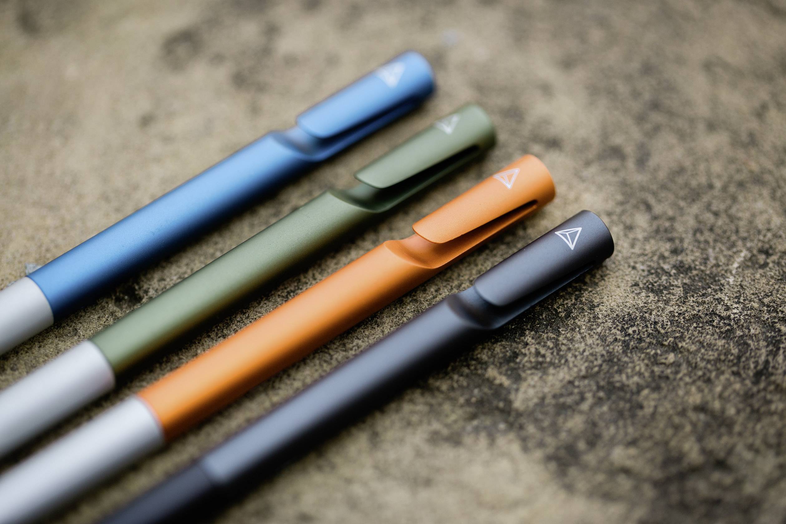 Vier kleurrijke pennen in blauw, groen, oranje en zwart liggen naast elkaar op een gestructureerd oppervlak.