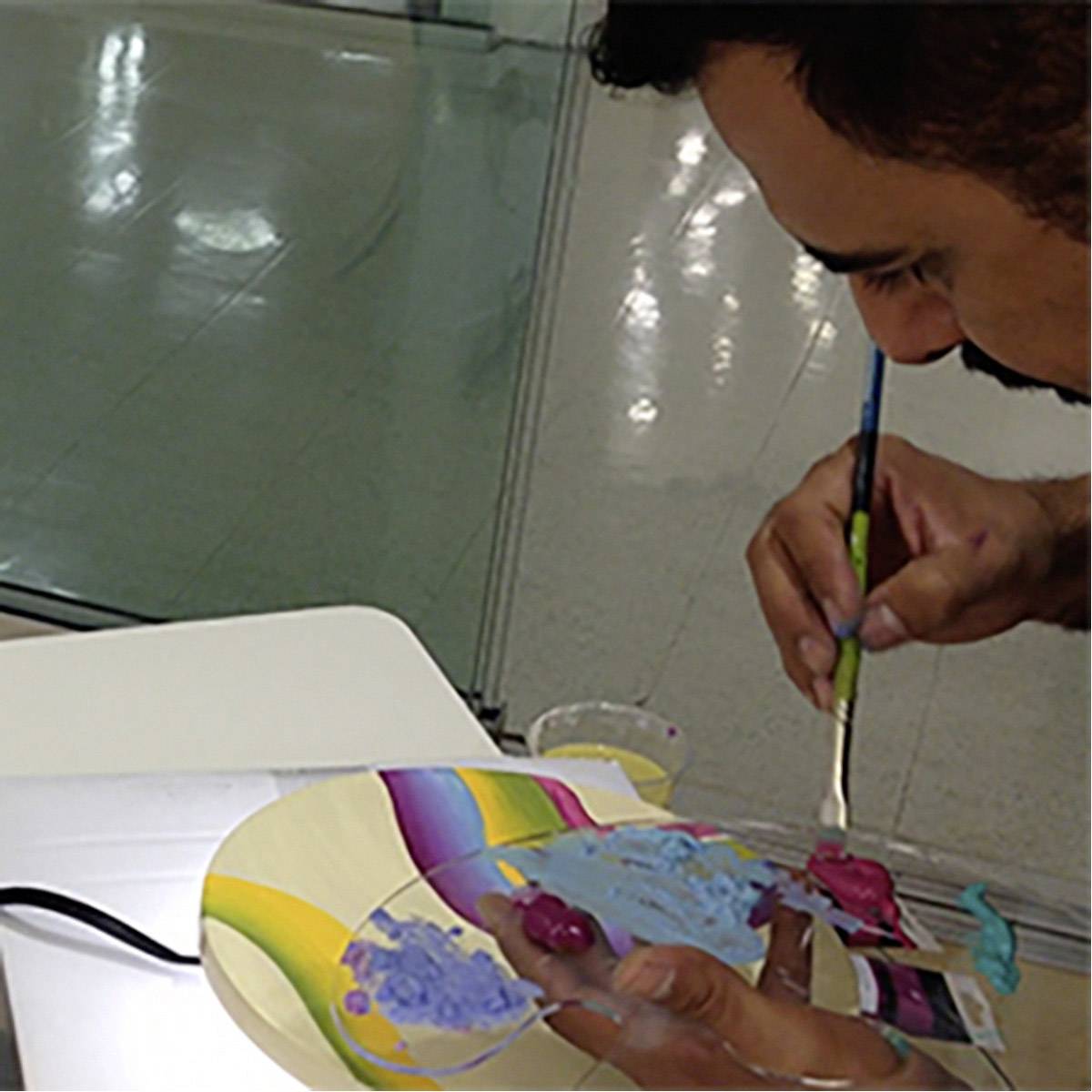 Een persoon schildert op een kleurenpalet met paarse en blauwe tinten, met een geconcentreerde houding in een lichte ruimte.