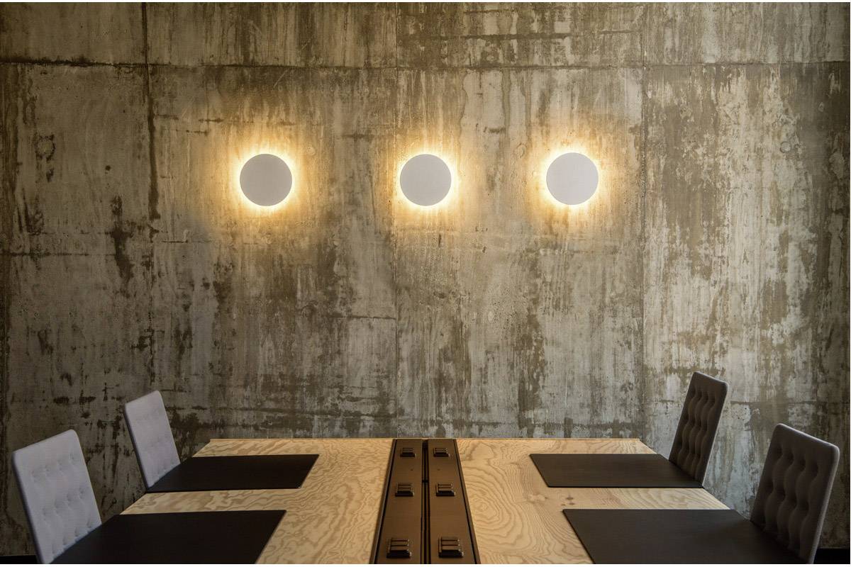 Een vergaderzaal met een rechthoekige houten tafel en zes stoelen, verlicht door drie ronde wandlampen aan een betonnen muur.