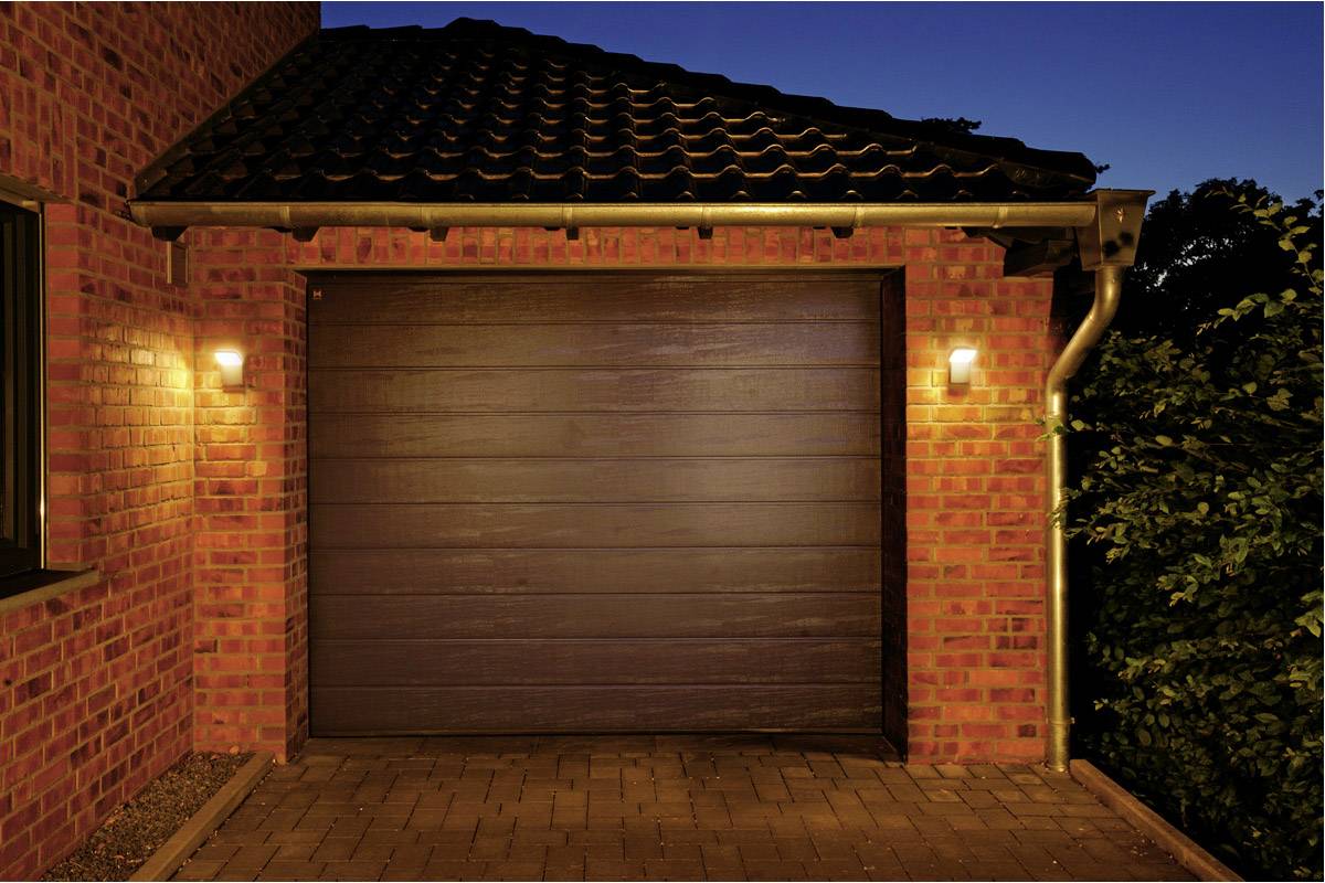Bakstenen huis bij schemering met een gesloten houten garagedeur, verlicht door twee buitenlampen.