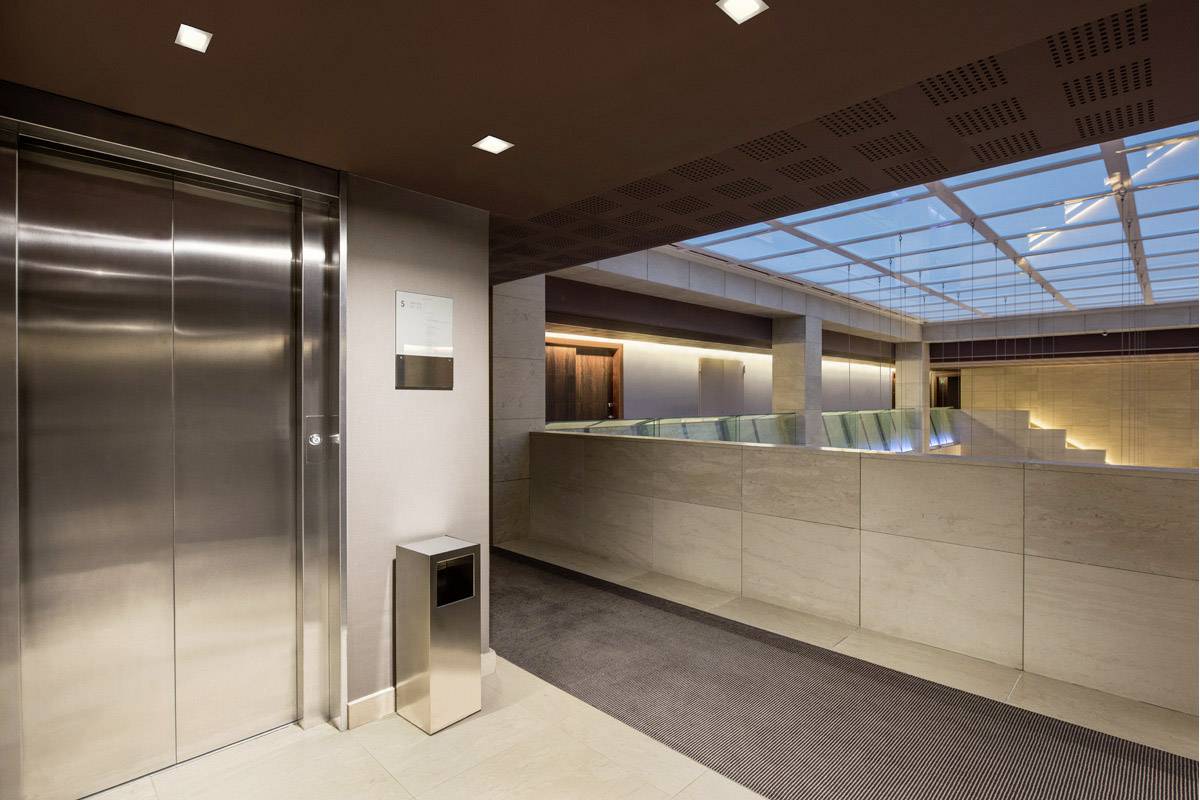 Een moderne lift in een gebouw met marmeren vloeren en een glazen dak, naast een vuilnisbak. Eenvoudige, strakke architectuur.
