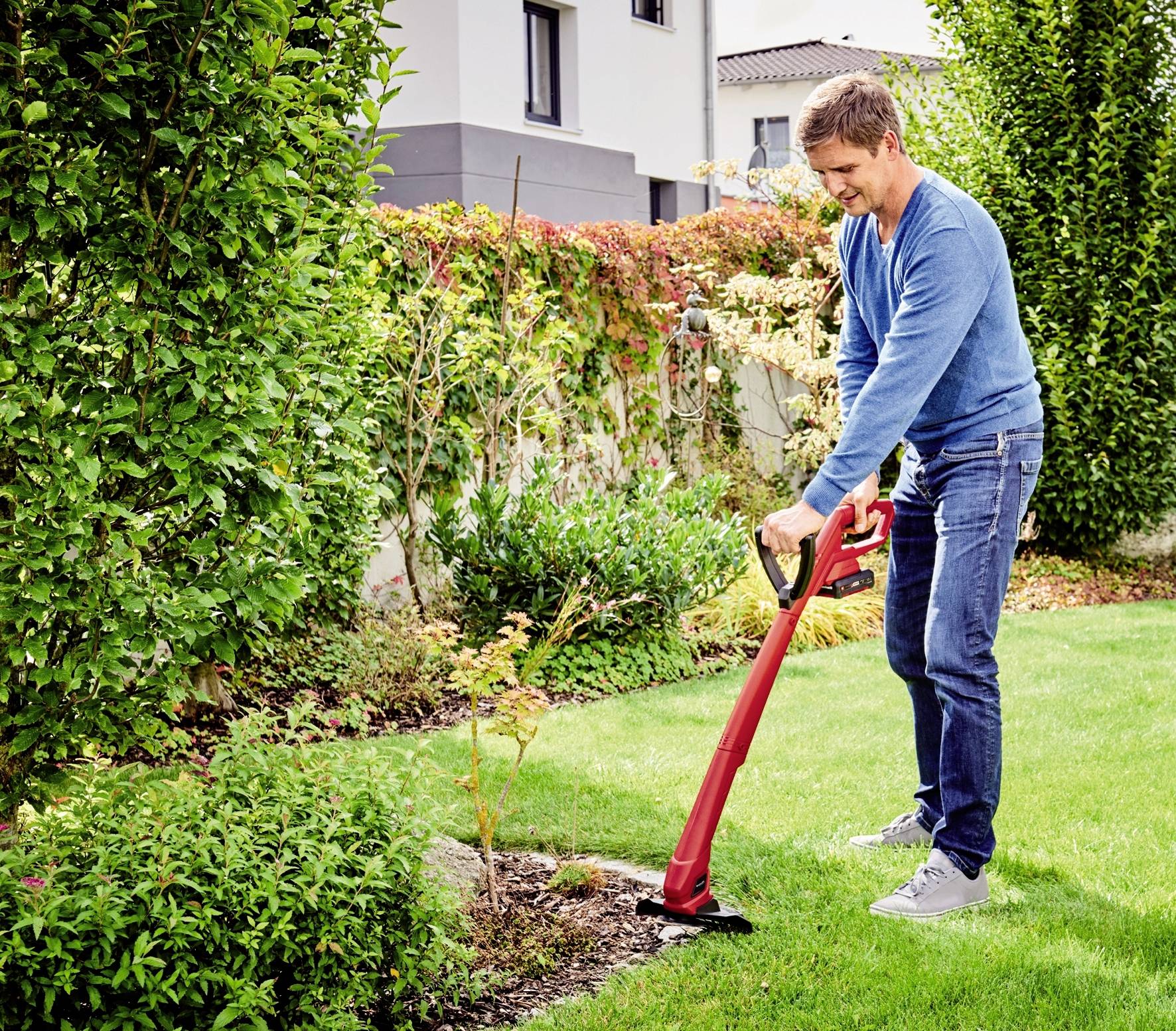 Een man in spijkerbroek en een blauwe trui gebruikt een rode gazonmaaier om het gras in een keurig verzorgde tuin te trimmen.