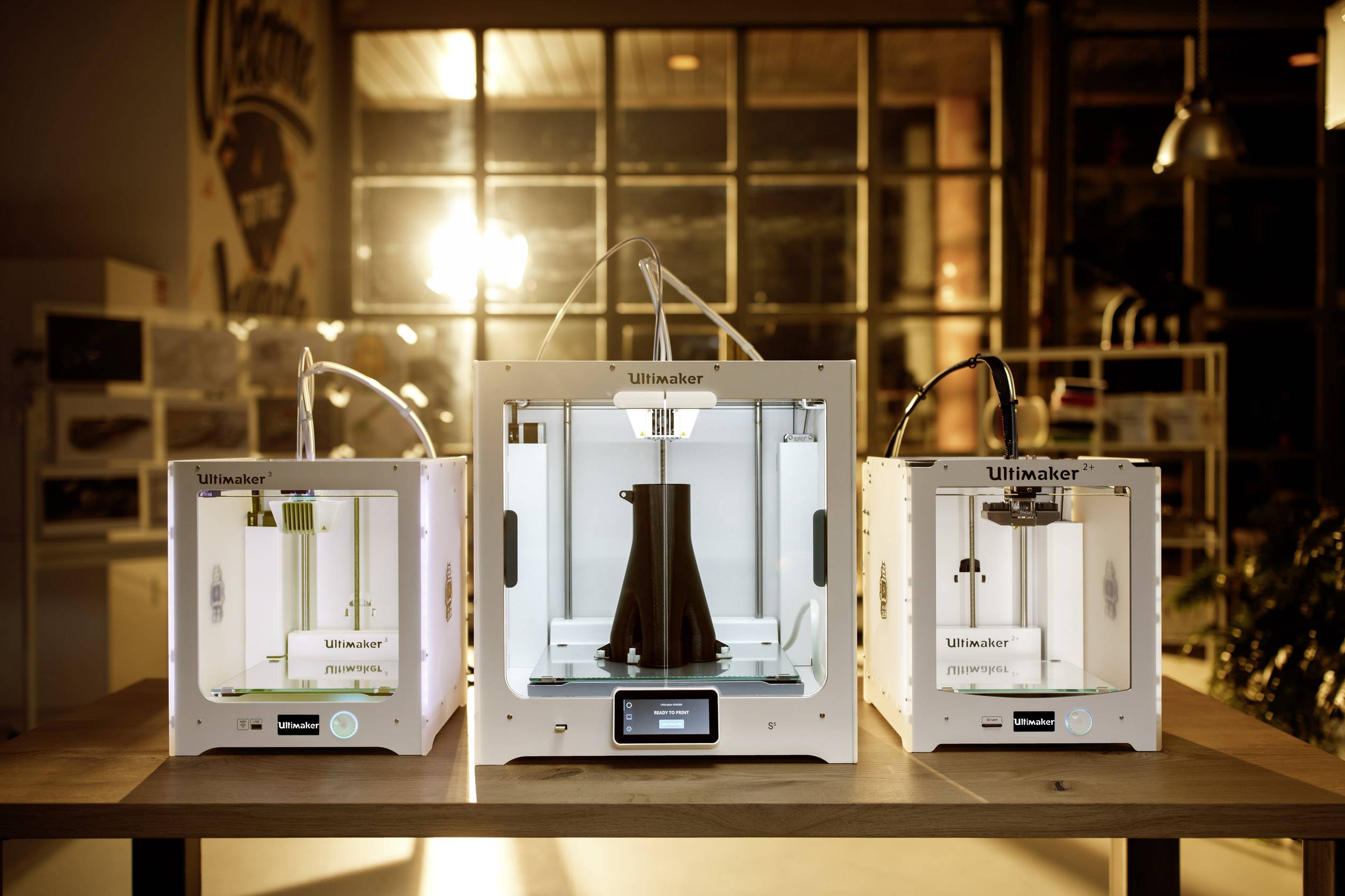 Drie 3D-printers op een tafel in een verlichte ruimte. De middelste printer is bezig een groot, zwart object te vervaardigen.