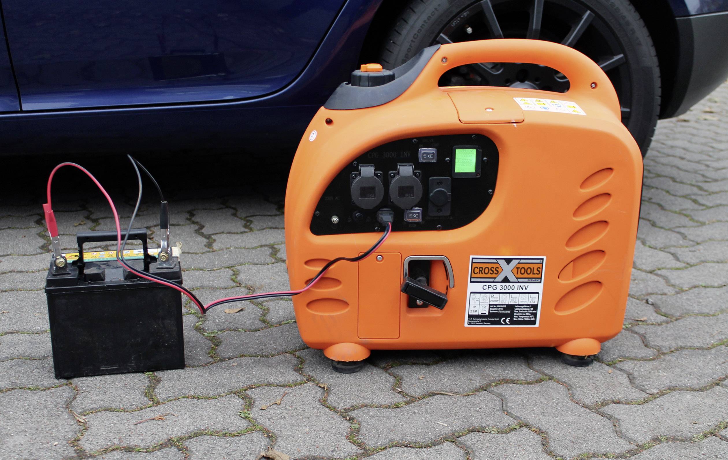 Een draagbare oranje generator is via kabels verbonden met een zwarte batterij. Beide apparaten staan op bestrating.