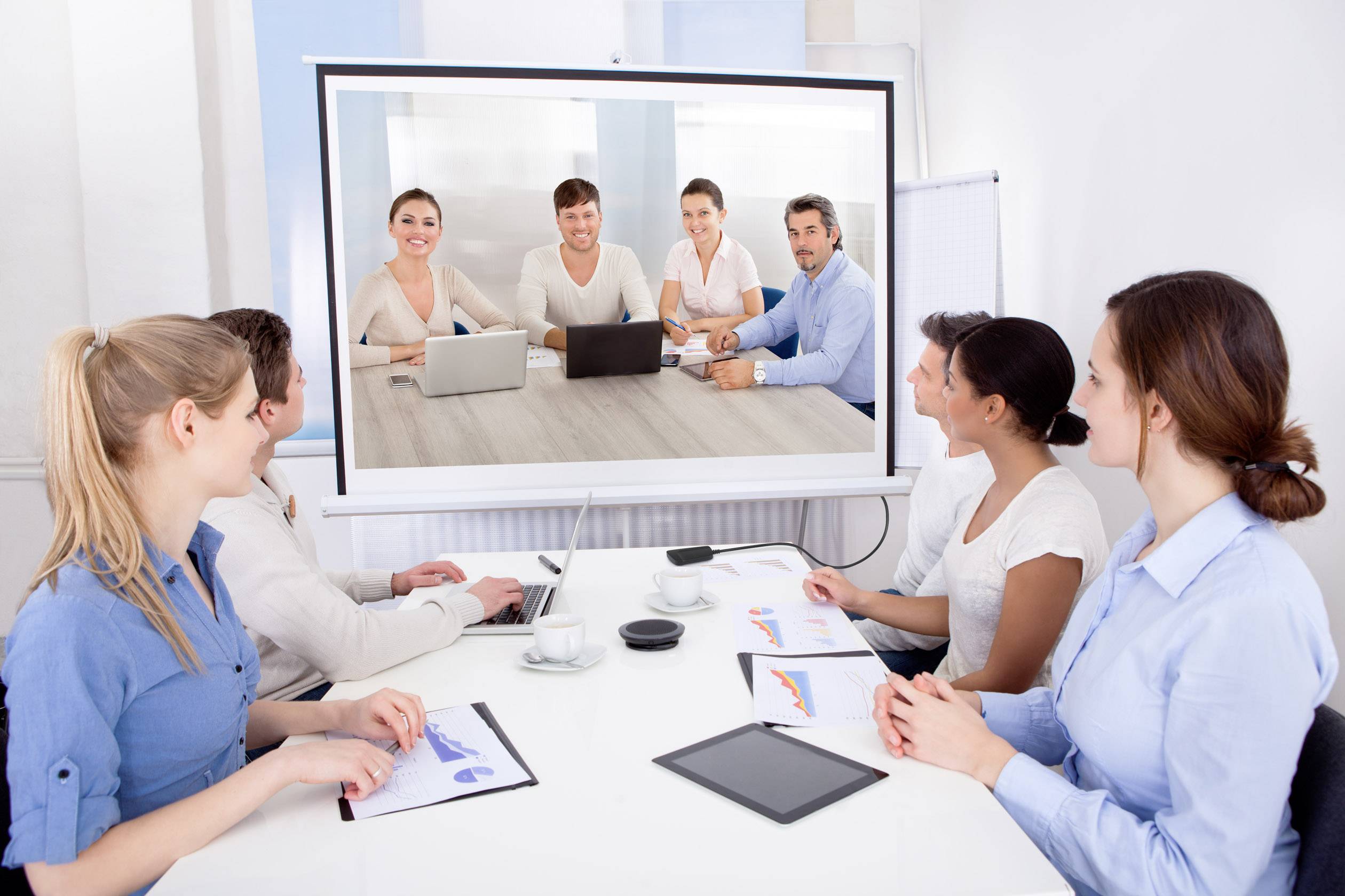 Zes personen nemen deel aan een videoconferentie in een kantoor. Twee zitten aan tafel met laptops, terwijl vier anderen op een scherm te zien zijn.