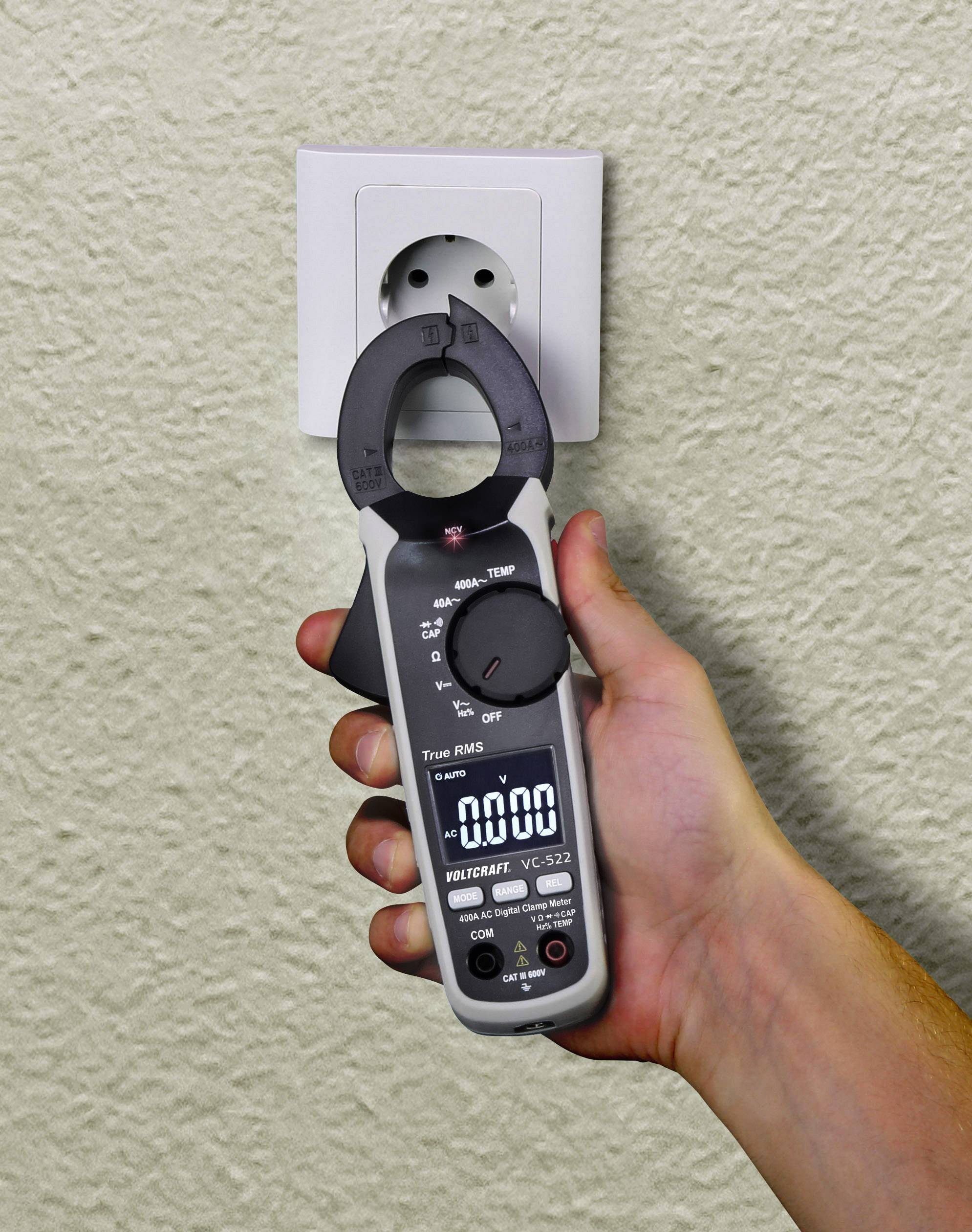 Een hand houdt een spanningsmeters voor een stopcontact. Het meetapparaat toont "0.00" aan. Er wordt spanning gemeten.