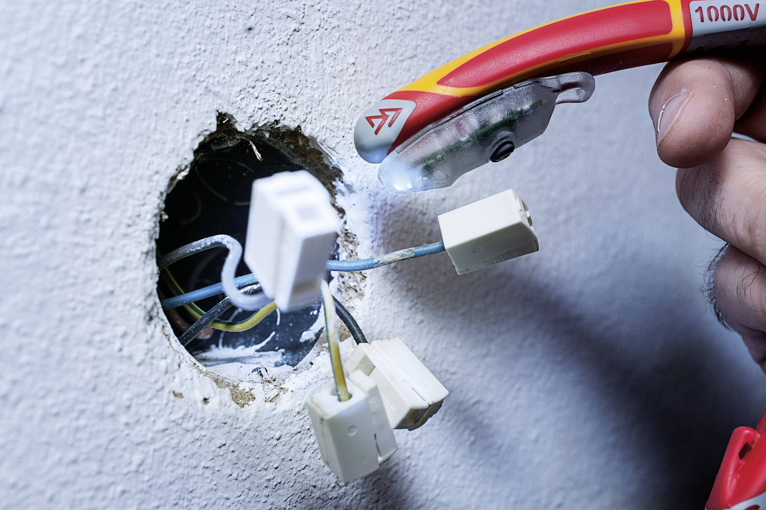 Een hand houdt een spanningzoeker vast bij open elektrische kabels die uit een rond gat in de muur steken.