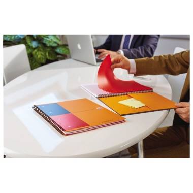 Twee personen bij een vergadering aan tafel. Eén werkt op een laptop, de andere bladert door een notitieboek. Notitieblokken liggen op tafel.