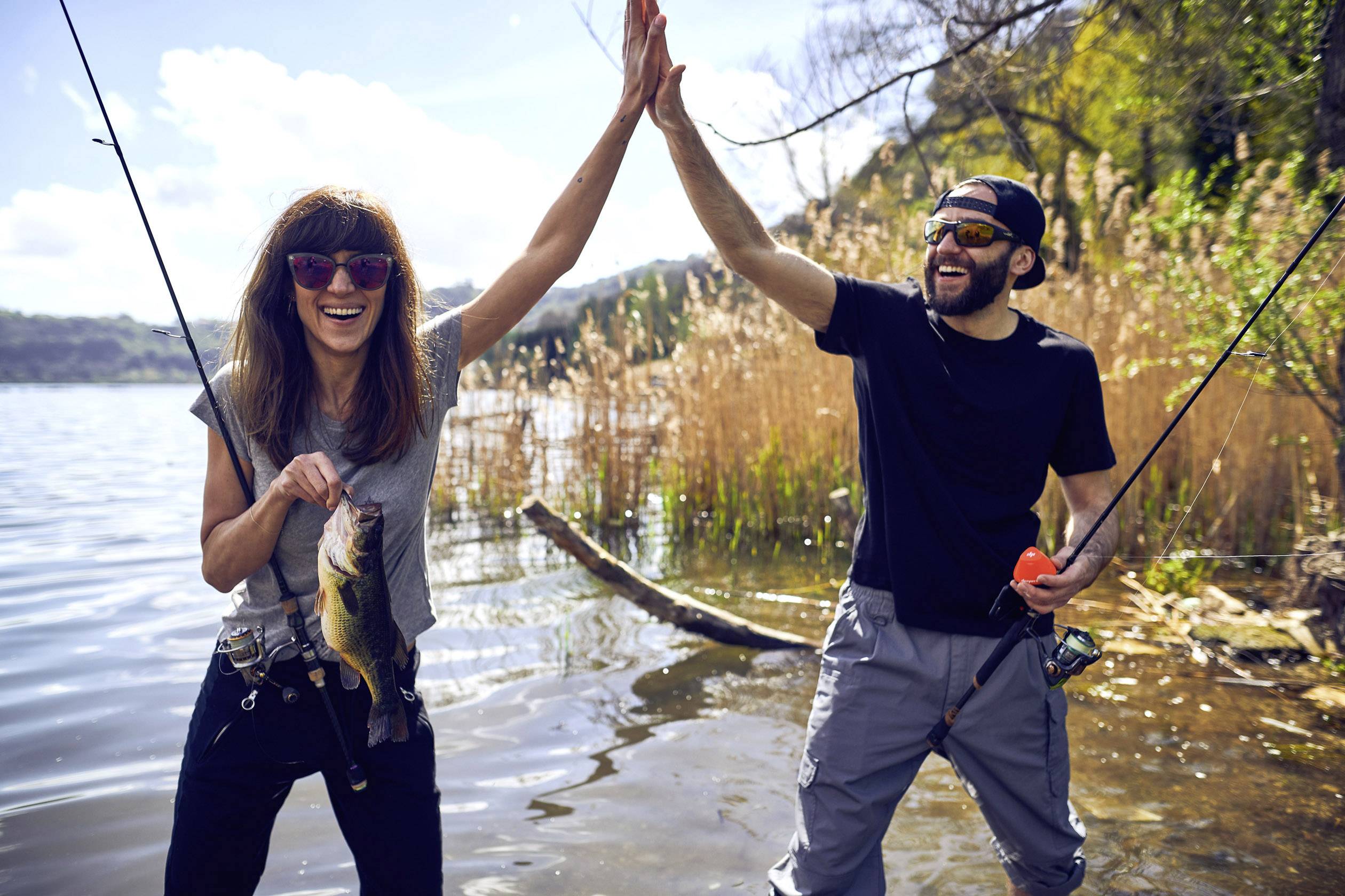 Een vrouw en een man staan aan de oever van een meer. Ze houdt een vis vast en ze geven elkaar een high-five. Op de achtergrond zijn riet en bomen te zien.