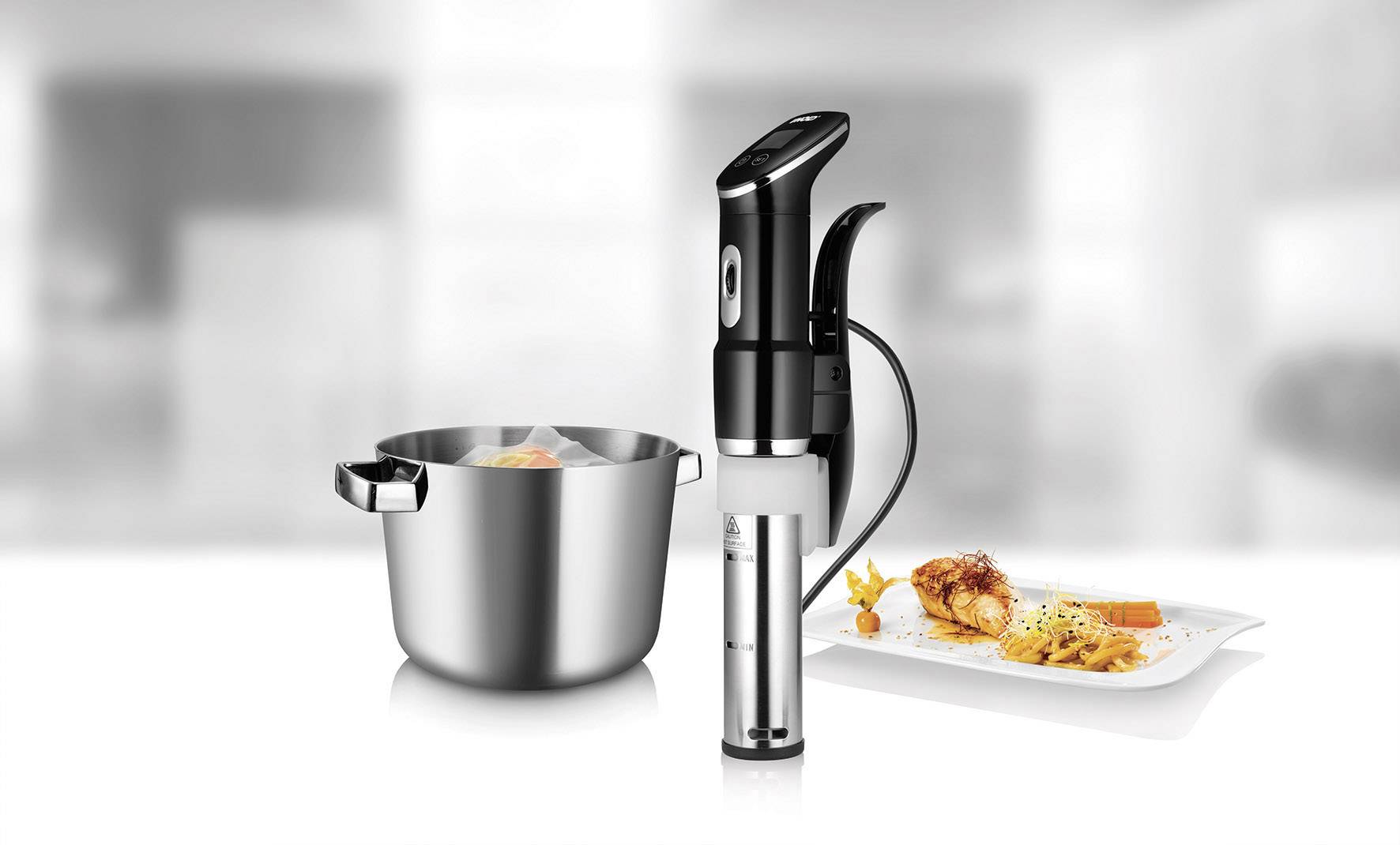 Sous-vide-apparaat naast een pan met ingrediënten en een bord met geserveerd vlees en groenten. Geplaatst in een moderne keuken.