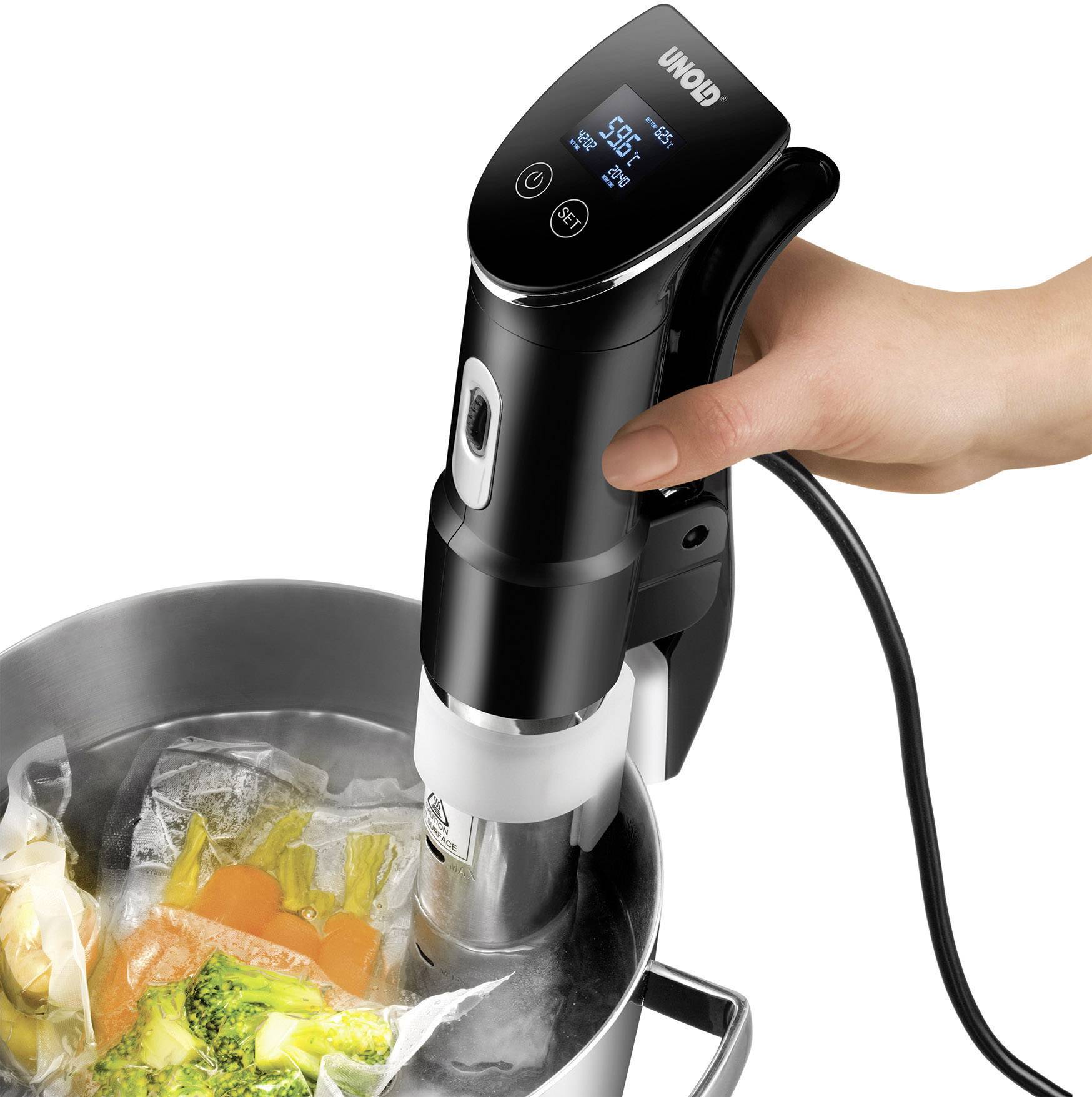 Een hand houdt een sous-vide-staaf vast die in een pan met vacuüm verpakte groentezakjes wordt gedompeld. Digitaal display toont temperatuur.