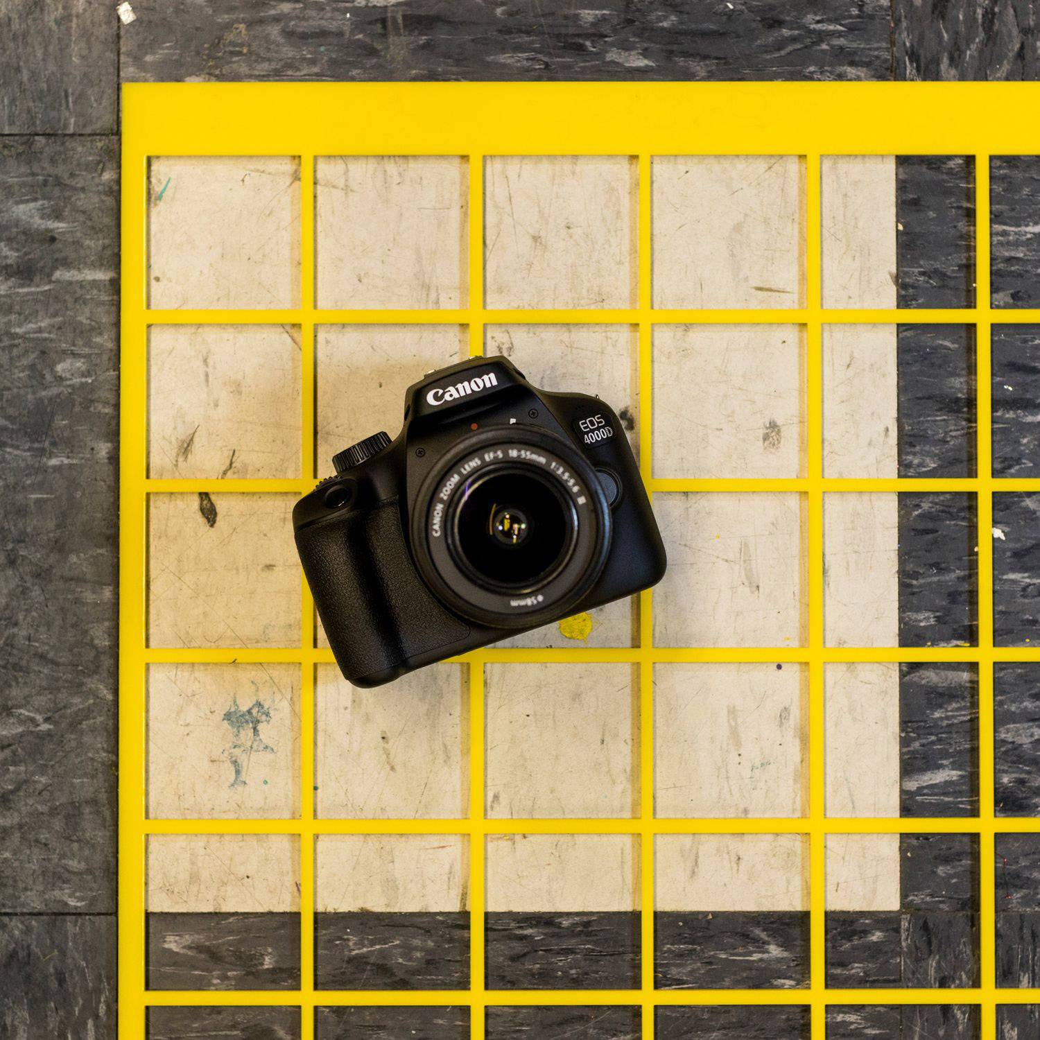 Een zwarte camera ligt op een geruit vloerpatroon met gele lijnen. De camera is van bovenaf gefotografeerd.