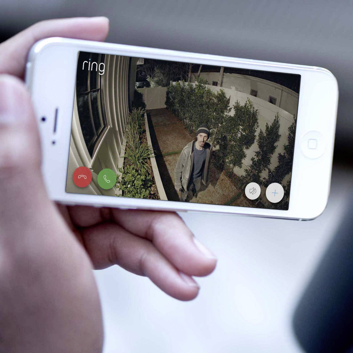 Een persoon houdt een smartphone vast met een video-bewaking die een persoon voor een huis laat zien. Tekst 'Ring' verschijnt linksboven.