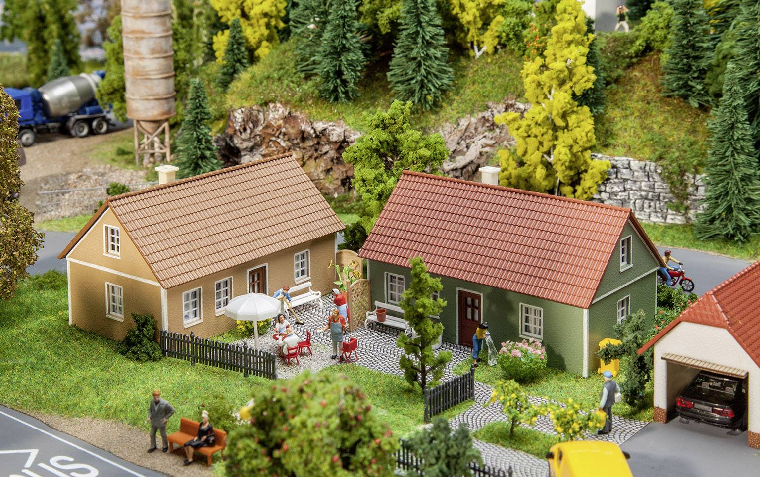 Miniatuurmakette van een dorp met twee huizen, figuren zittend en lopend, bomen op de achtergrond, een weergave van een landelijk tafereel.