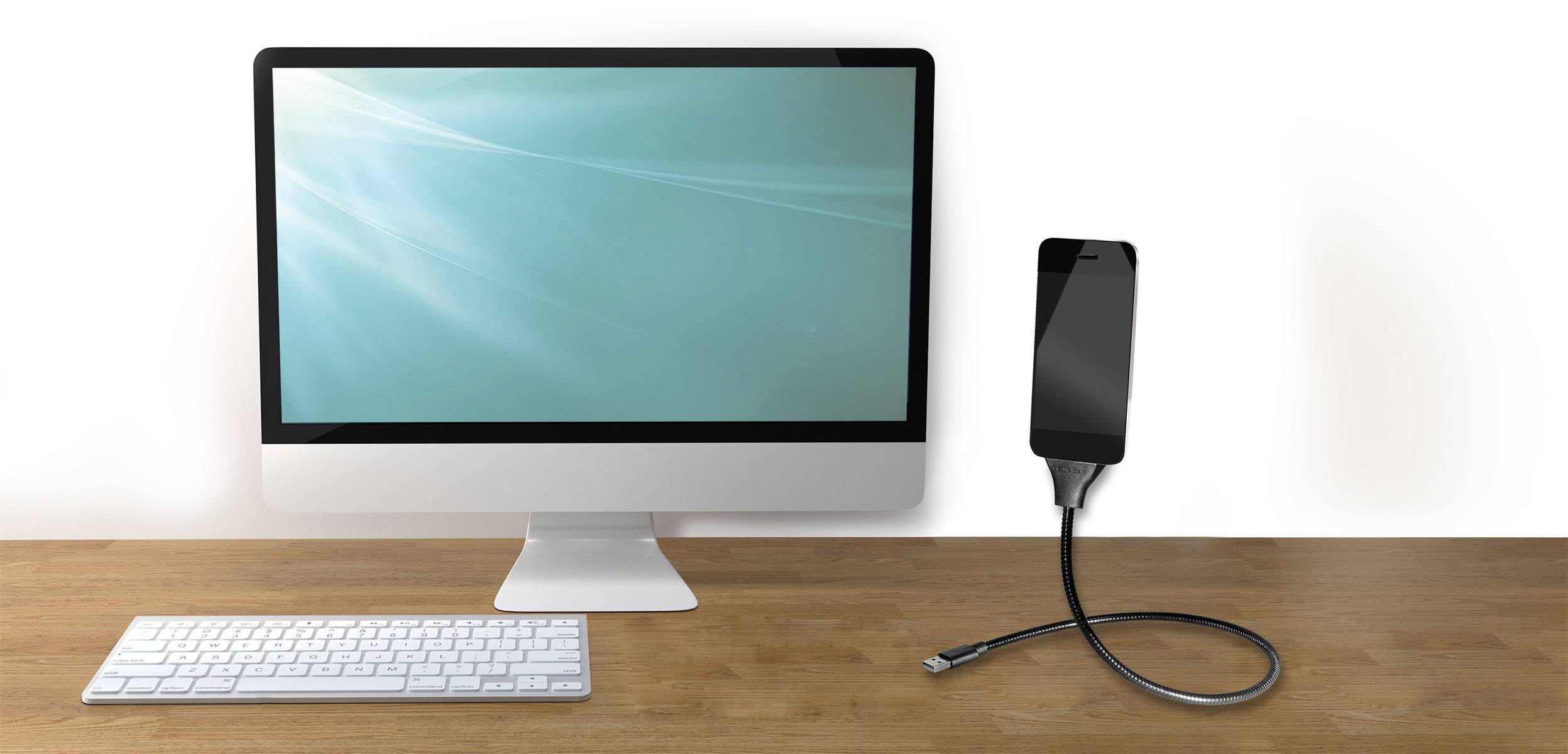 Een computerscherm en een toetsenbord op een tafel, verbonden met een smartphone via een oplaadkabel.